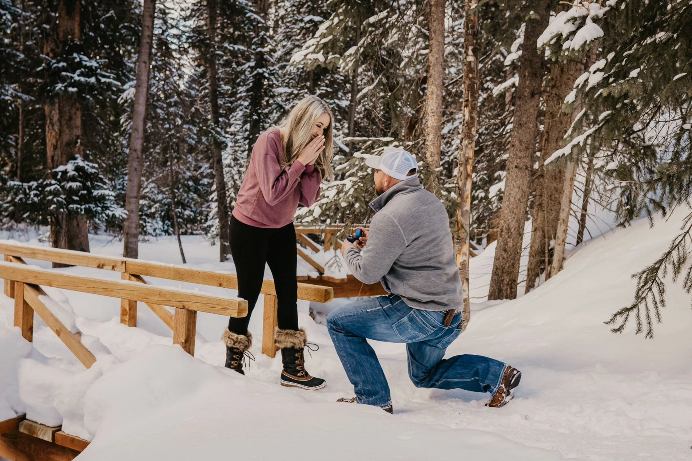 A man proposes to a woman on a snowy trail in a forest, with the woman covering her mouth in surprise and the man kneeling with a ring box.
