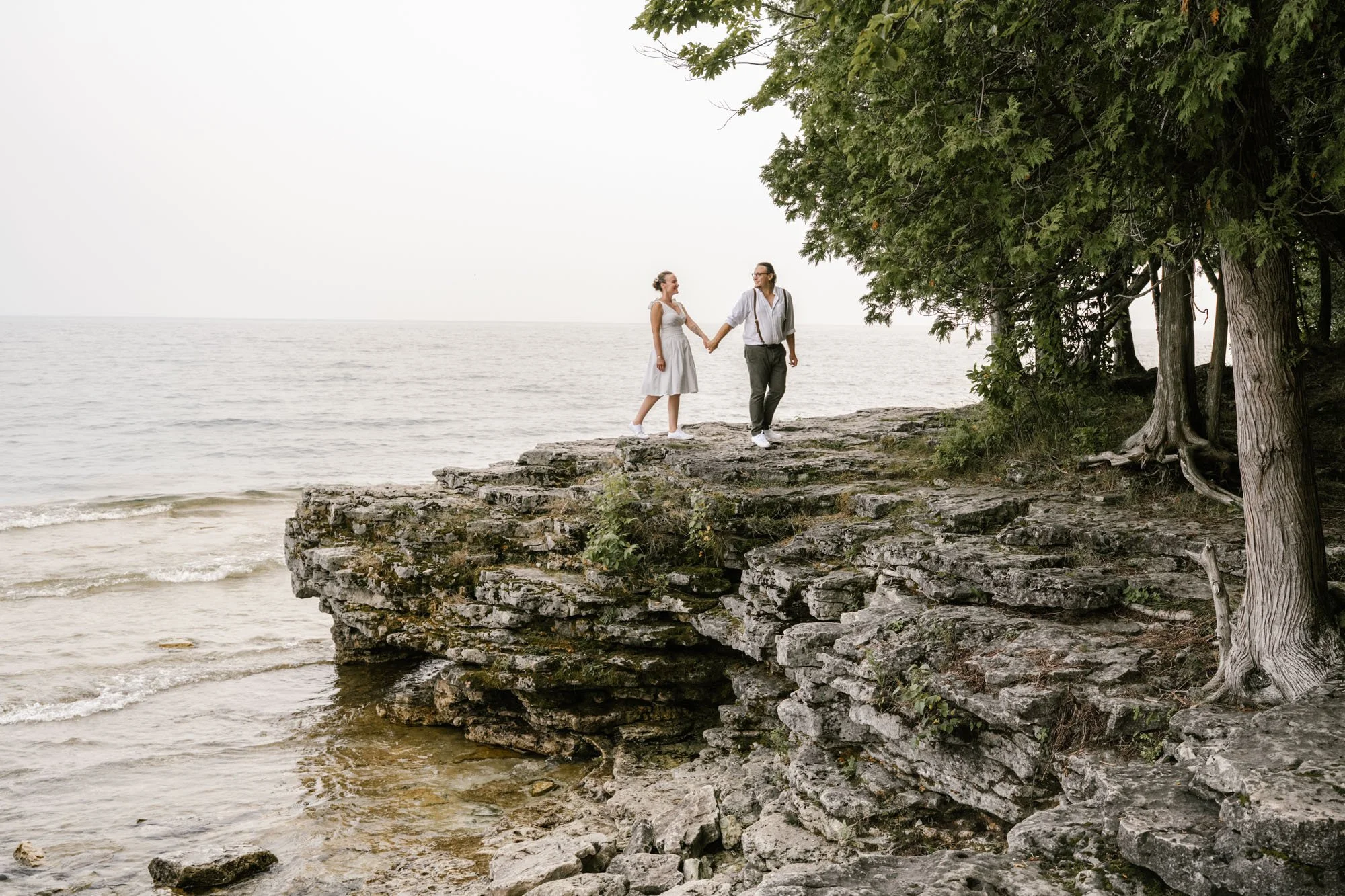 A couple in vintage wedding day clothing holding hands on rocky seaside cliff with trees nearby and choppy waves in the background.