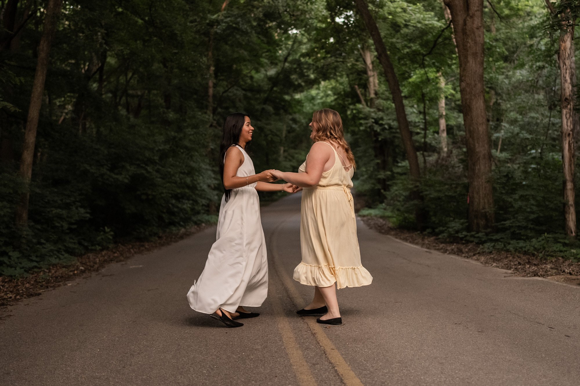 Two women in dresses holding hands and smiling at each other on an empty road surrounded by tall trees in a forest.