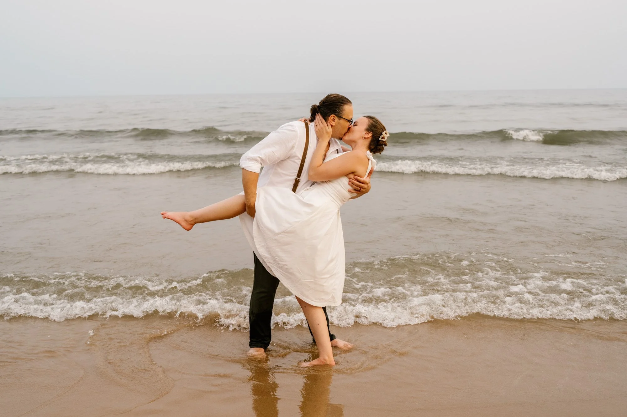 A couple in wedding attire sharing a kiss at the beach, with the groom holding the bride in his arms near the shoreline, ocean waves in the background.