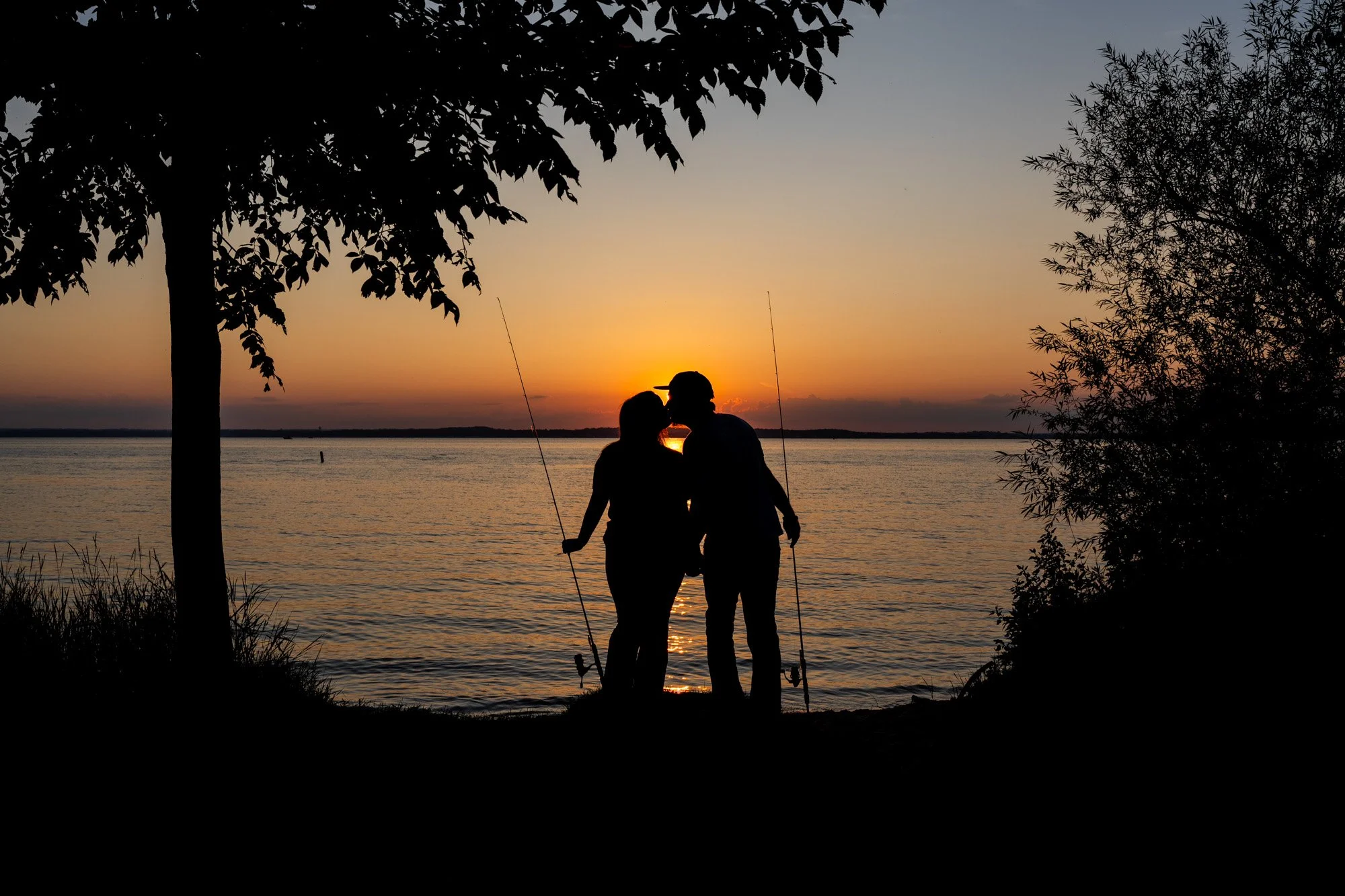 Silhouettes of a couple with fishing poles standing by the water at sunset, framed by trees.
