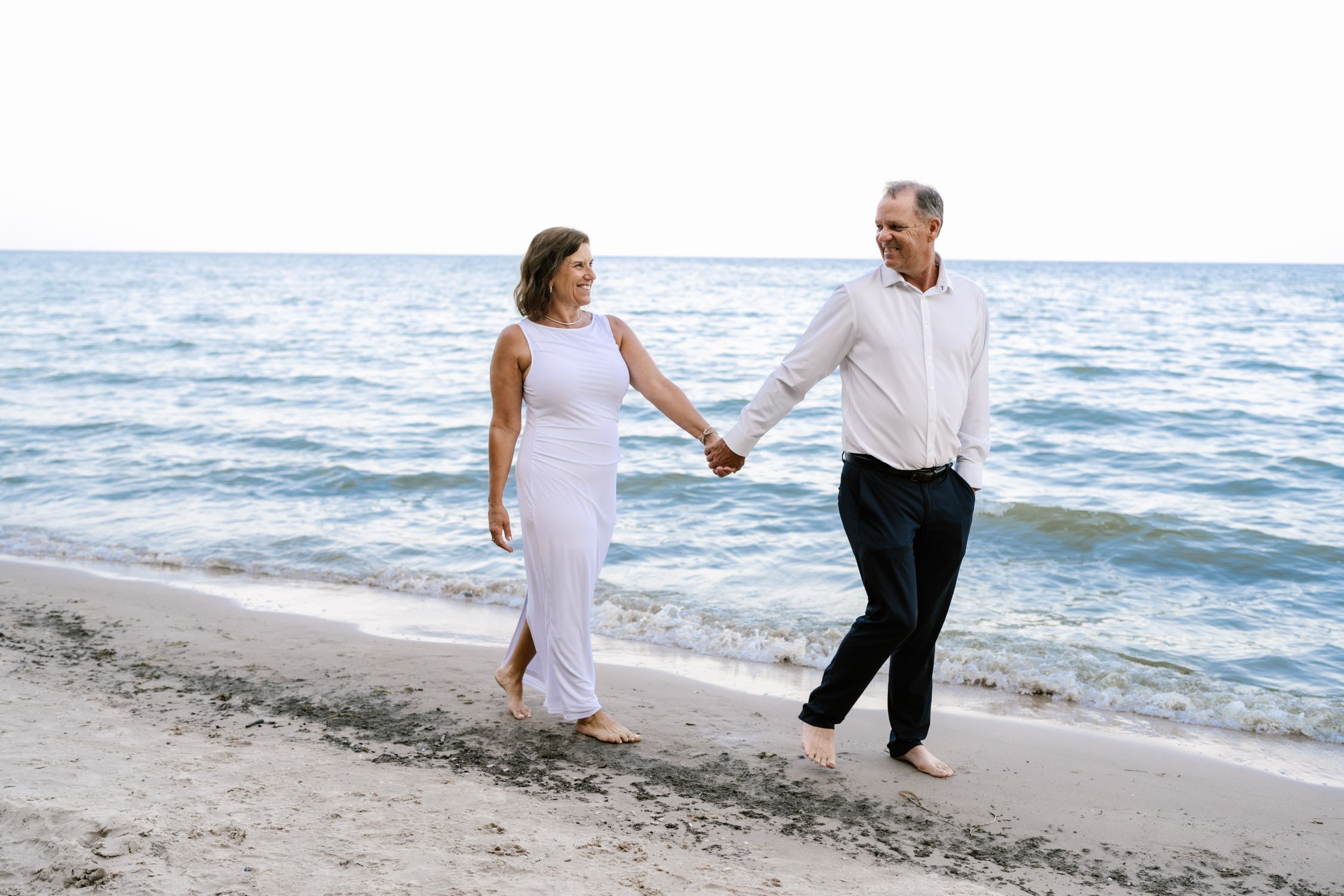 A middle-aged couple holding hands and walking barefoot along the shoreline of a beach, smiling at each other with the ocean in the background.