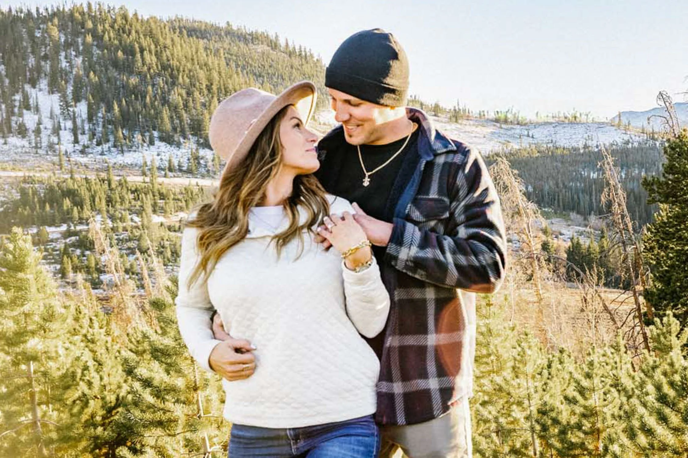 A woman and a man standing close together outdoors in a forested trail area with snow, trees, and blue sky, looking at each other lovingly.