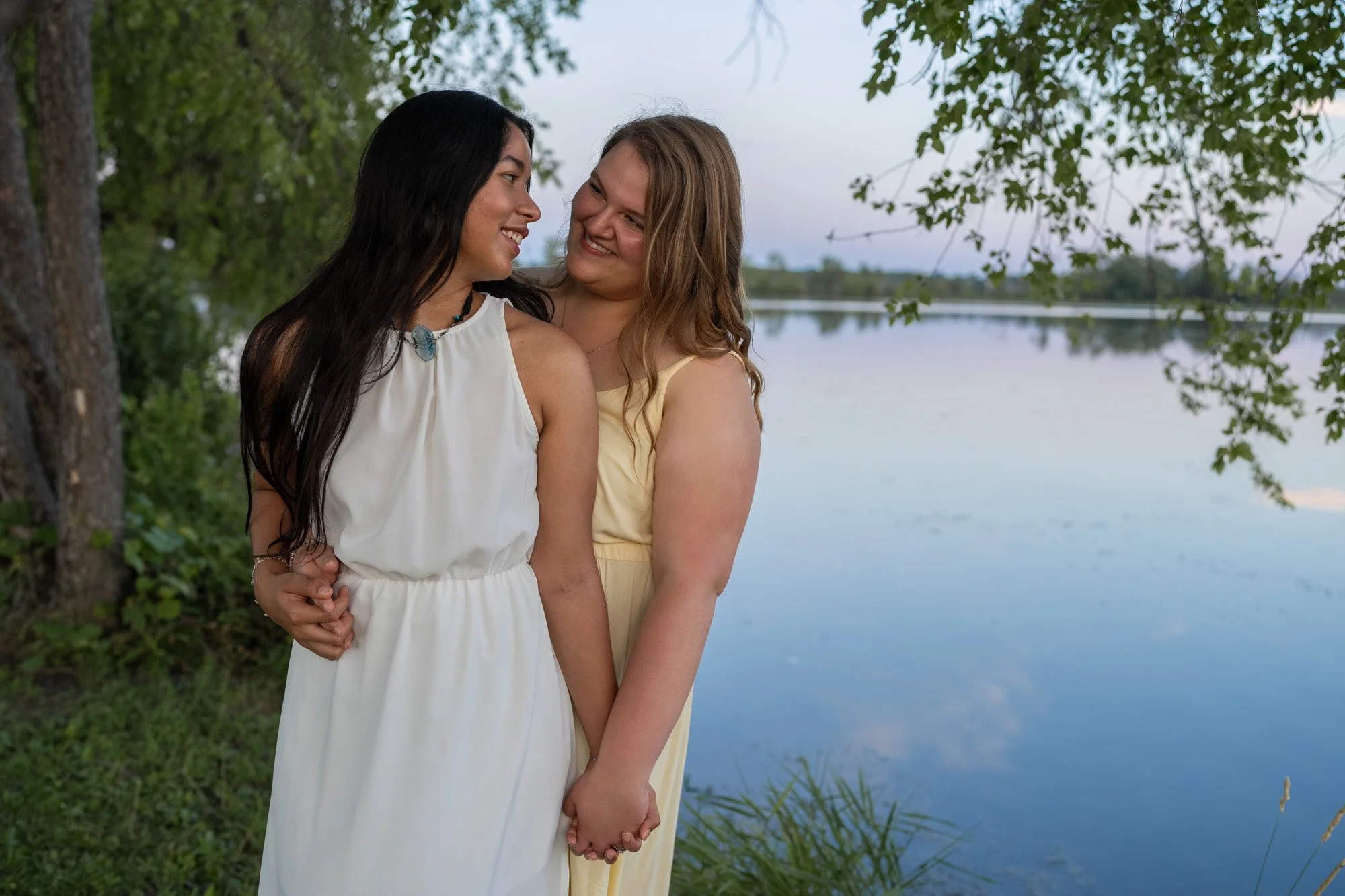Two women, one with black hair and the other with blonde hair, standing close and holding hands near a lake with trees in the background, smiling at each other.