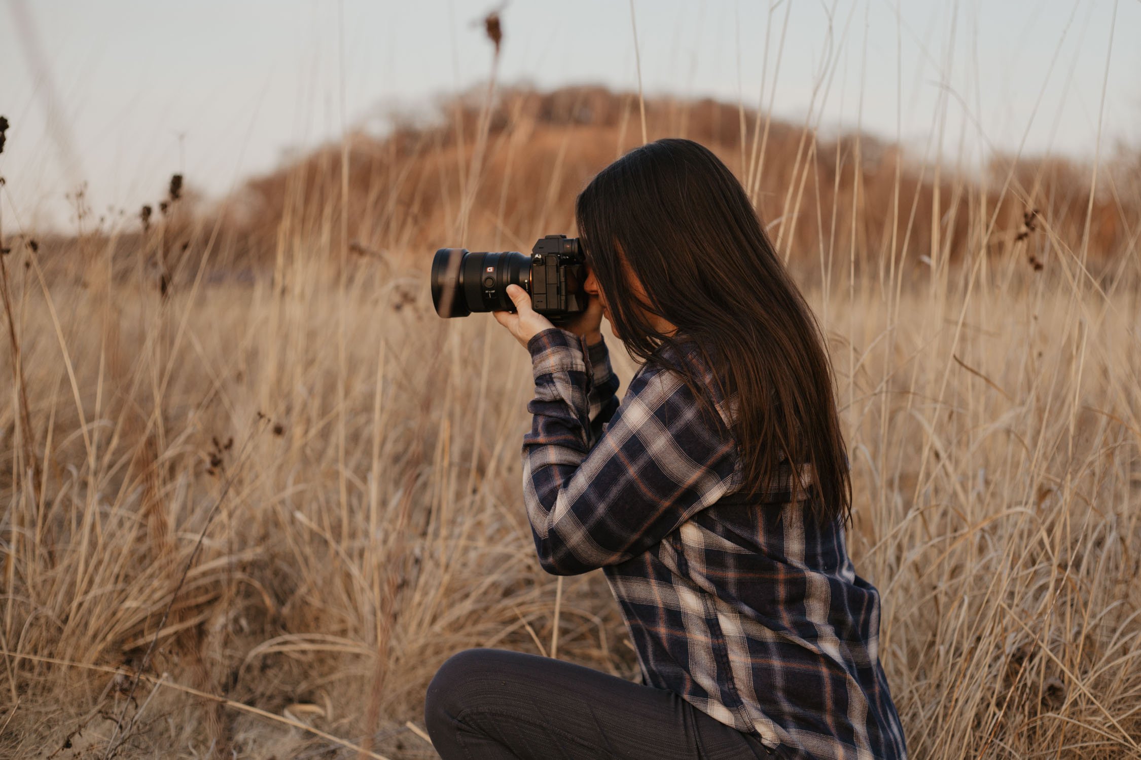 Woman with long dark hair wearing a plaid shirt taking photographs with a DSLR camera in a dry grassy field.