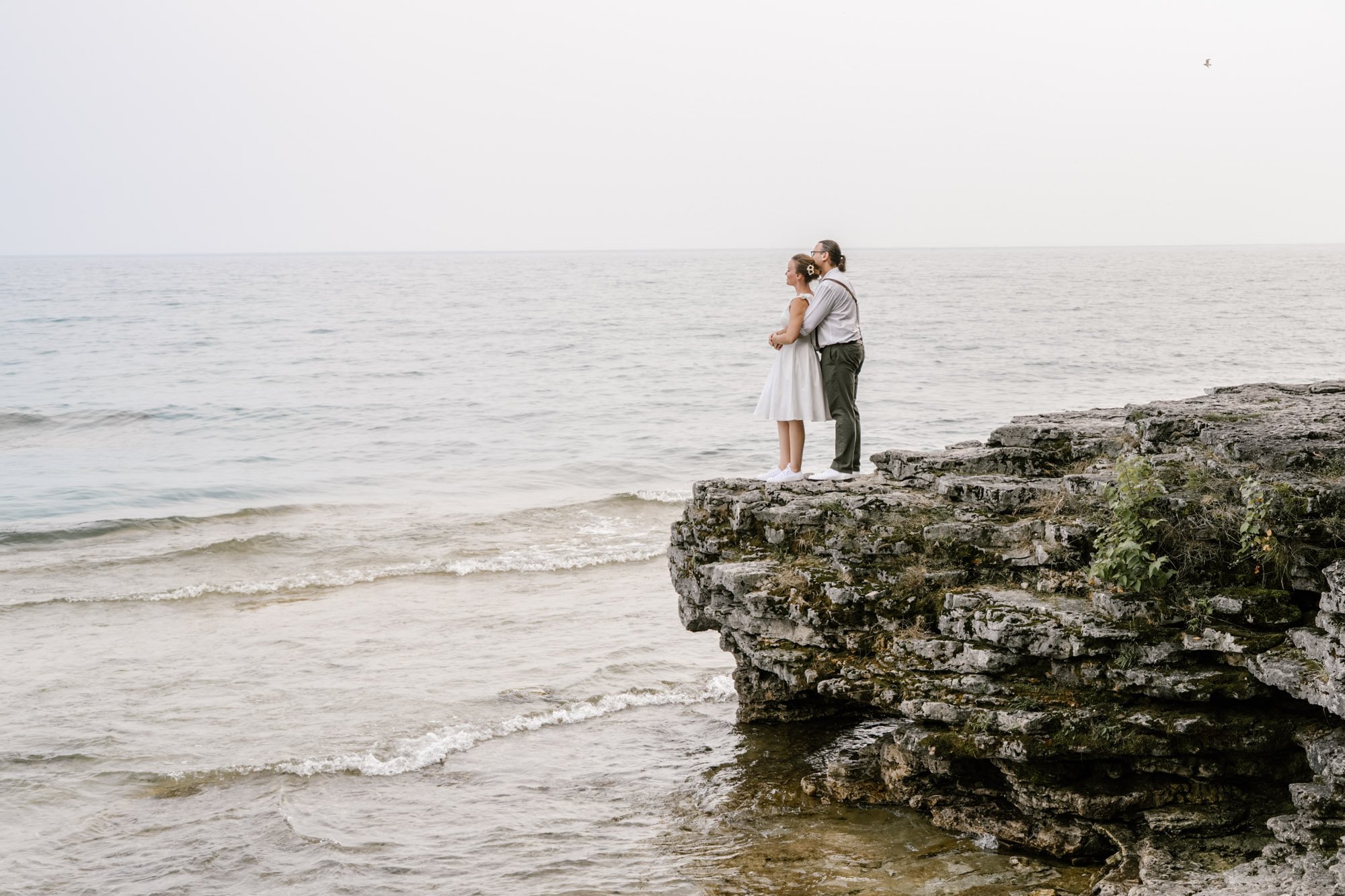 A couple stands on a rocky ledge overlooking the ocean, with the woman in a white dress and the man in a white shirt and gray trousers for their elopement day attire, embracing each other as they look out at the water during a cloudy day.