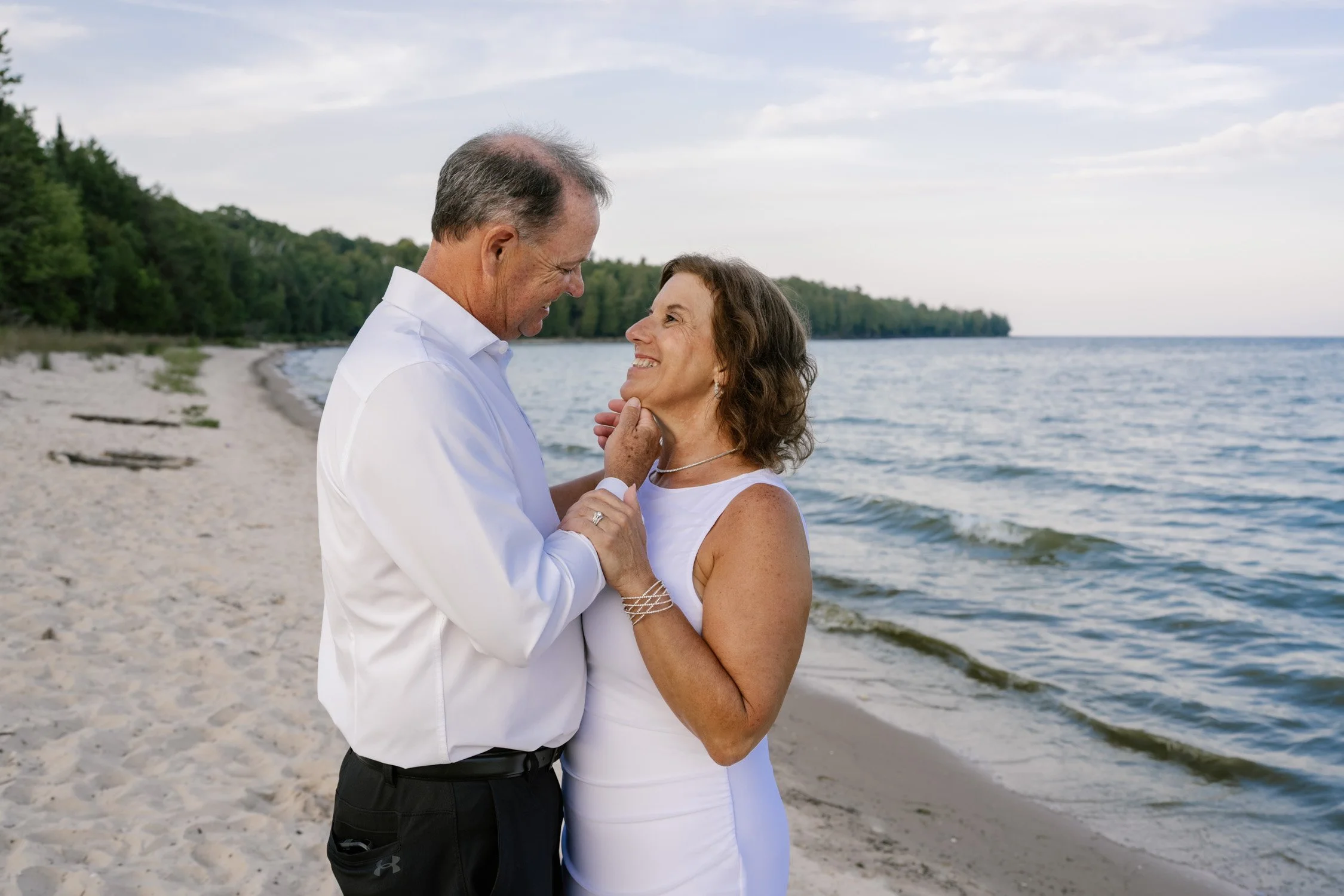 A man in a white shirt and a woman in a white dress on a beach, smiling and holding hands as they look at each other.