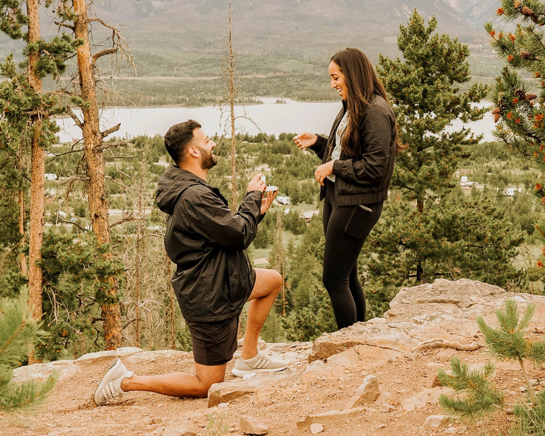 A man proposing marriage to a woman on a rocky outdoor trail with trees, a lake, and hills in the background.