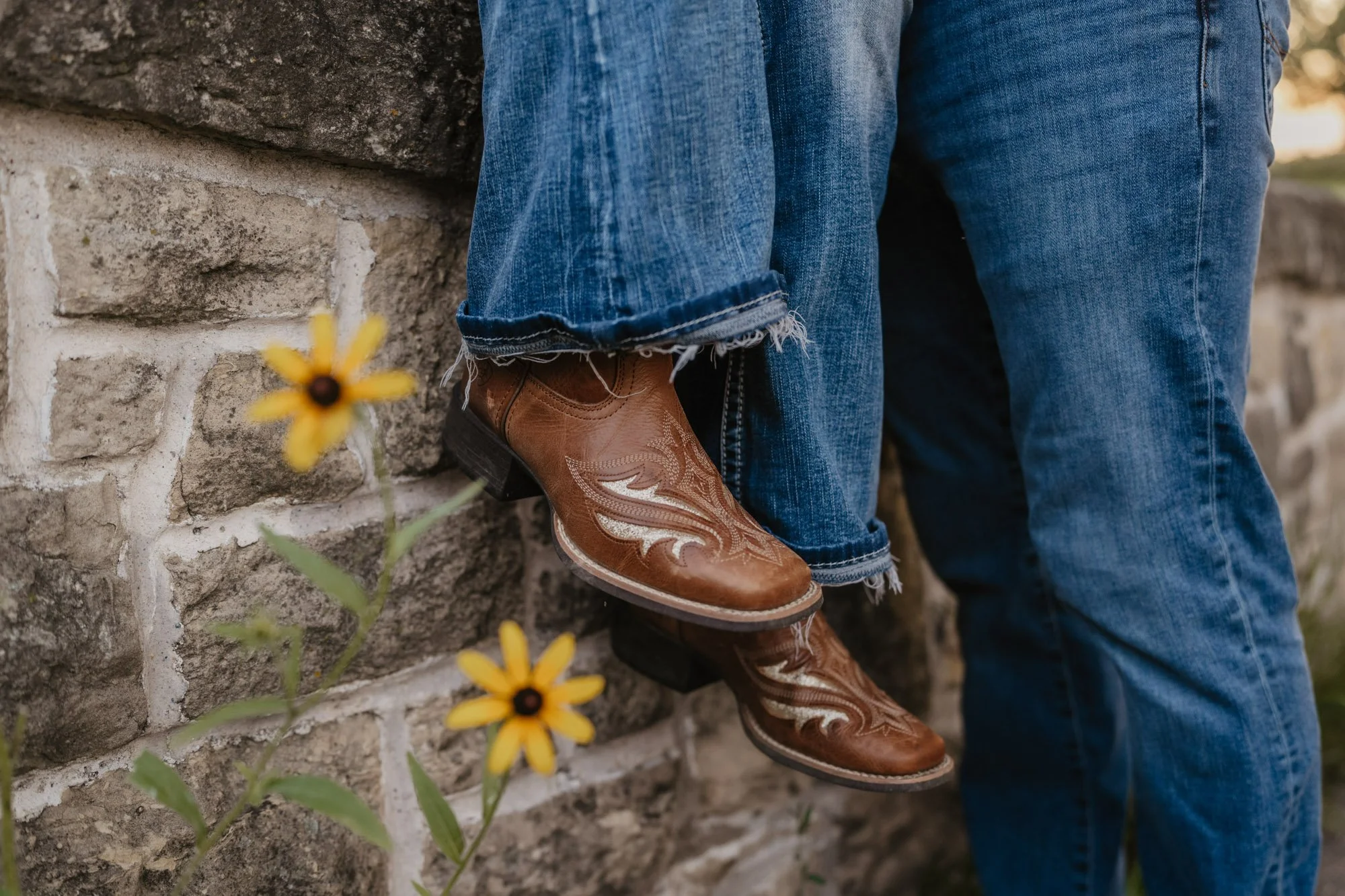 Close-up of brown cowboy boots with decorative stitching, worn by a person in distressed blue jeans with frayed hems, leaning against a stone wall with yellow flowers in the foreground.