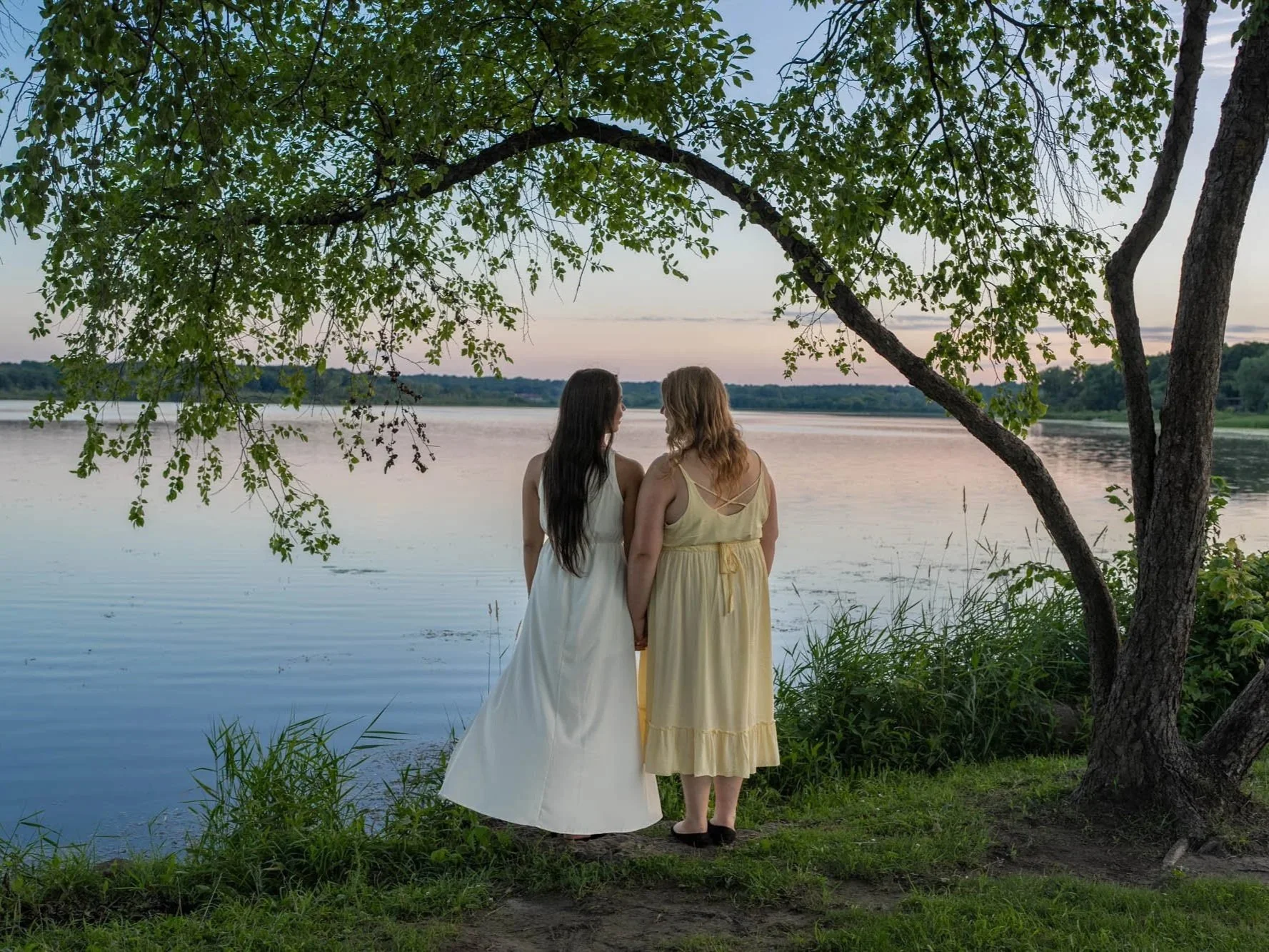 Two women in dresses stand hand in hand by a peaceful lake at sunset, under a tree with green leaves during their engagement session.