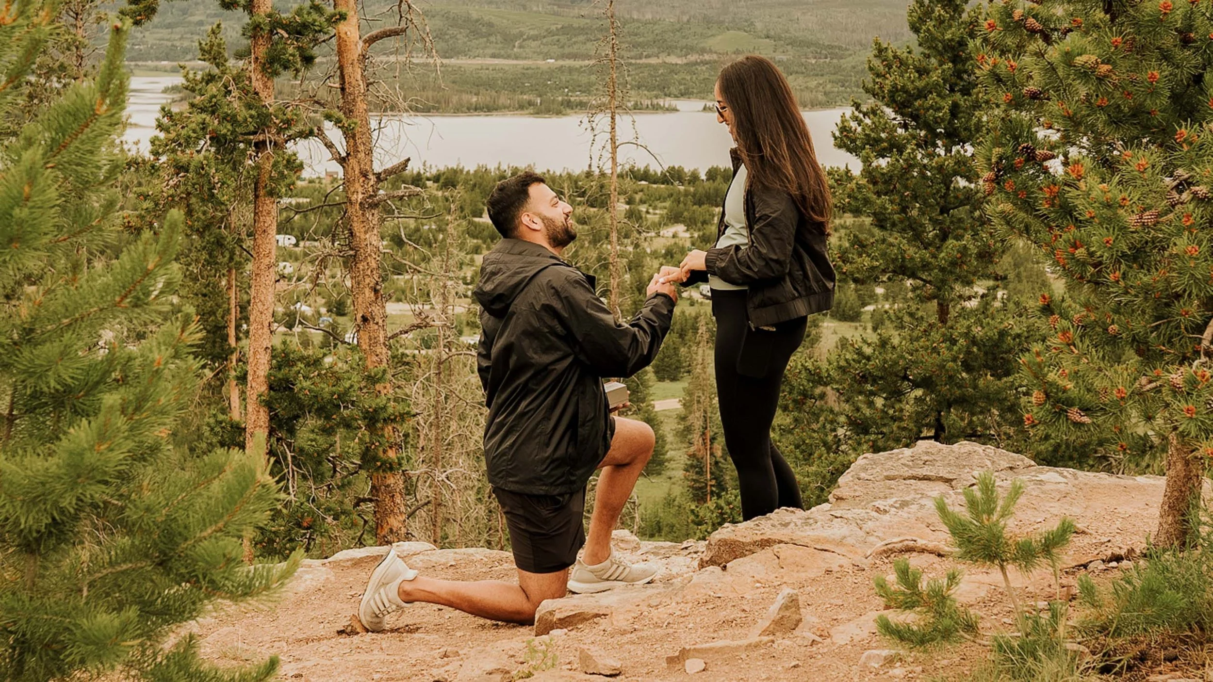 A man kneeling on one knee proposing to a woman on a rocky overlook surrounded by pine trees, with a lake and forest in the background.