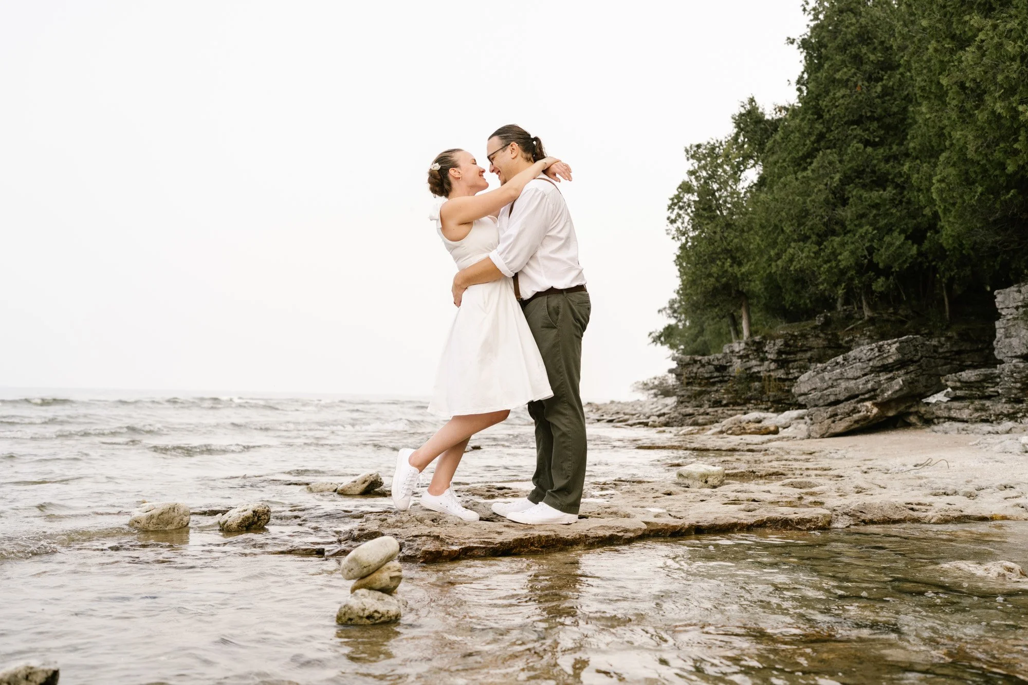 A couple embraces and smiles on a rocky beach with water and trees in the background just after they said their vows.