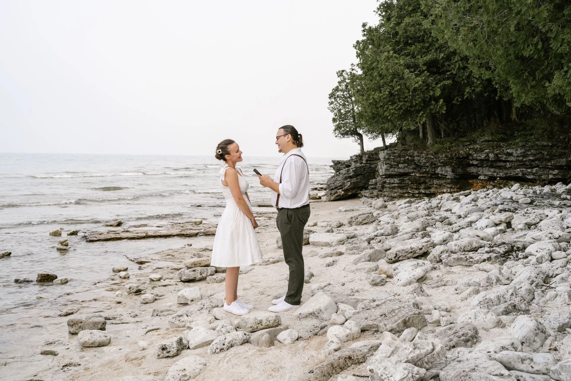 A couple stands on a rocky beach, facing each other, with the woman in a white dress and the man in dark trousers and a white shirt. They are smiling at each other while he reads his vows.
