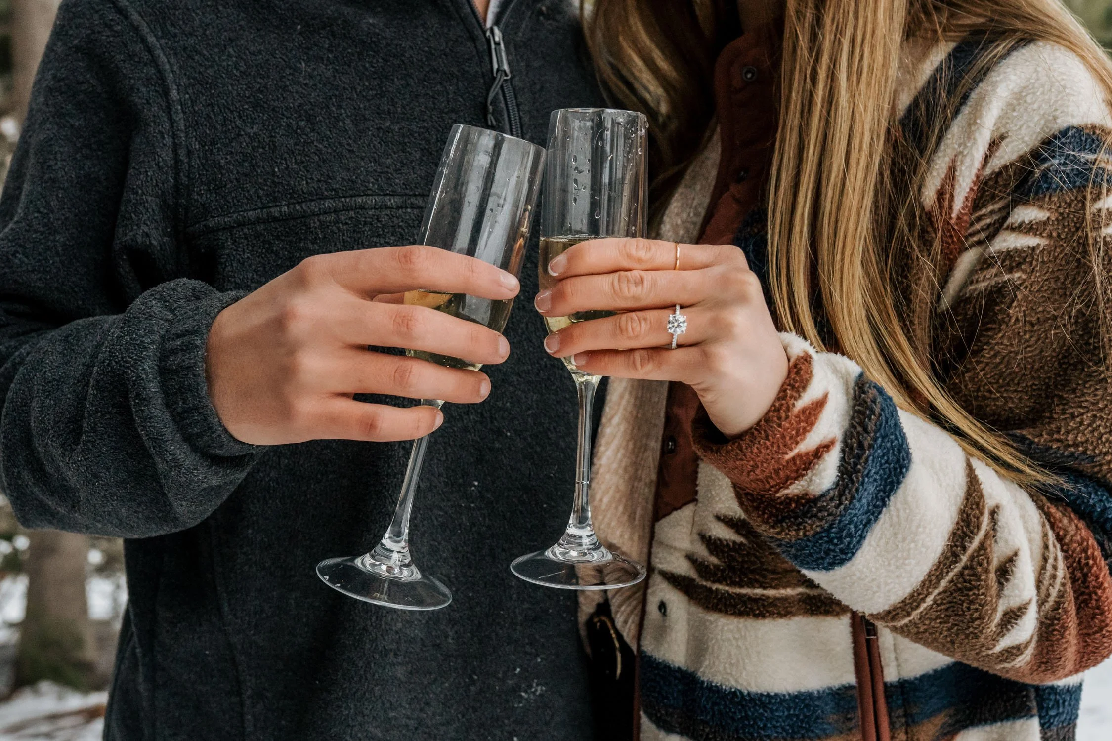 Two people clinking champagne glasses outdoors featuring an engagement ring.