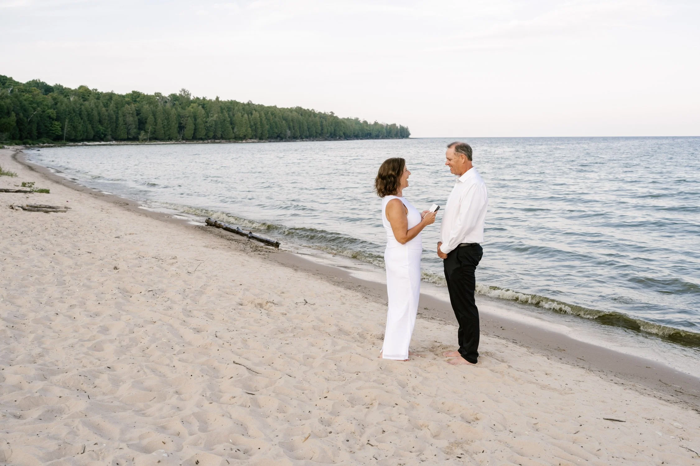 A couple standing on a sandy beach facing each other, with the woman holding a phone and the man barefoot, near the ocean.