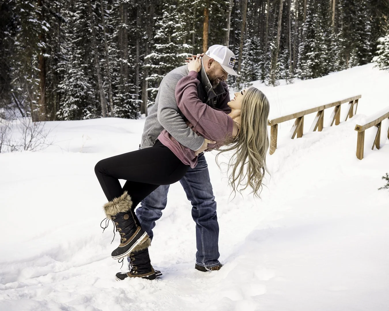A couple dancing in the snow in a forested winter landscape, with the man dipping the woman, both smiling and enjoying the moment.