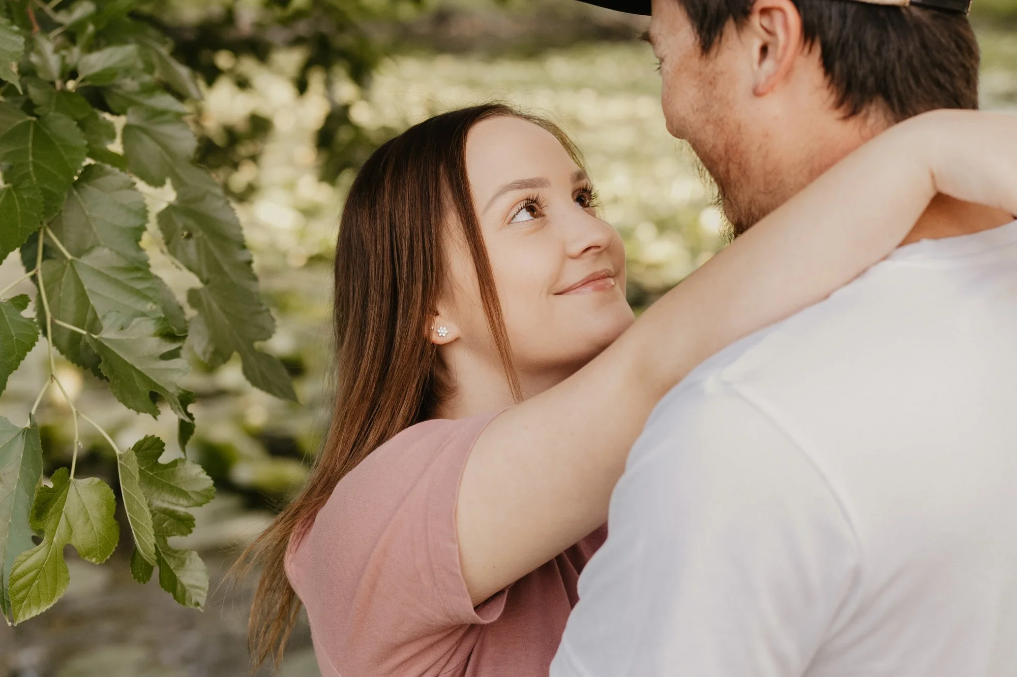 A young woman and a man embracing outdoors among green foliage, looking into each other's eyes.