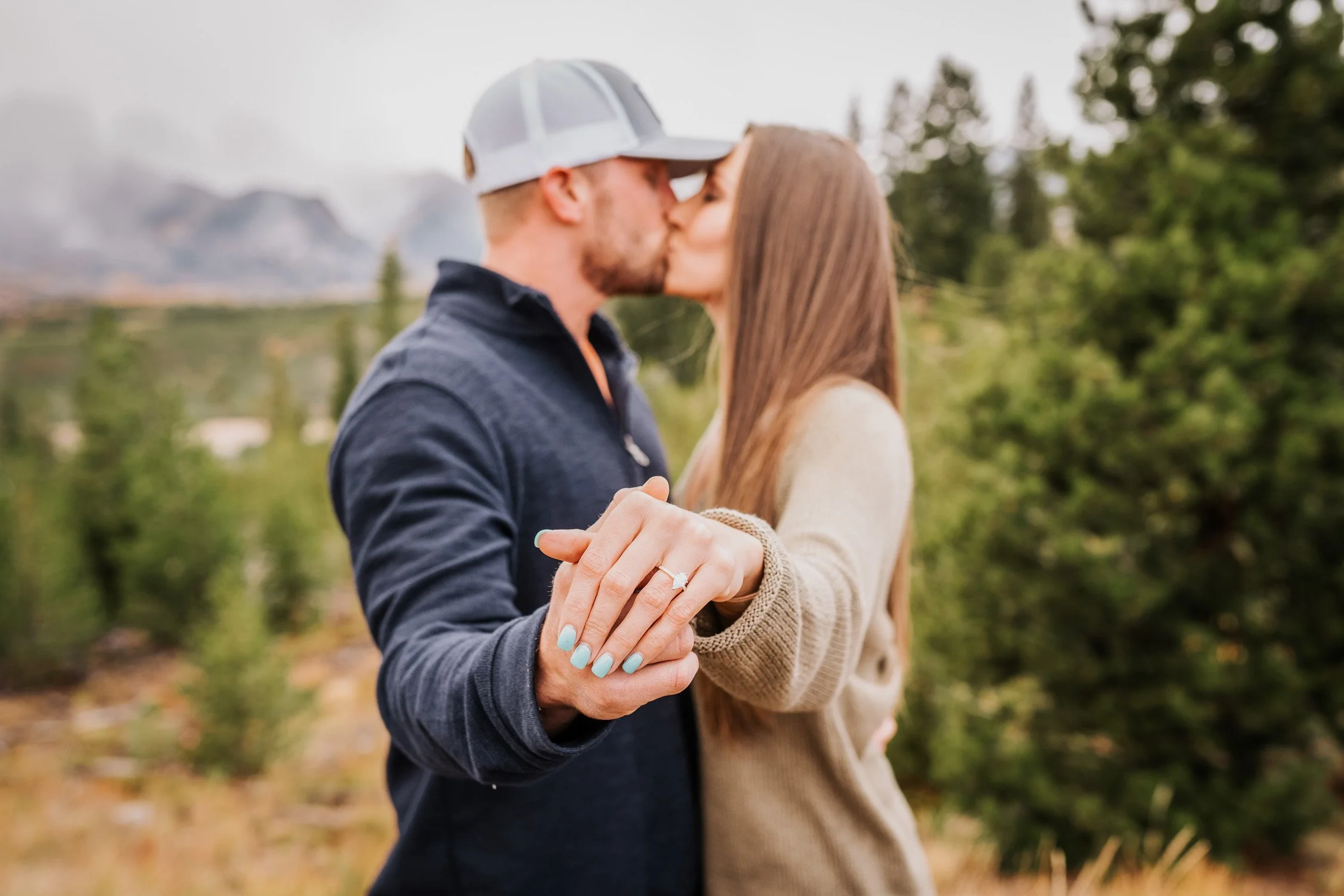 A couple kissing outdoors in a forested area, holding hands with an engagement ring visible.