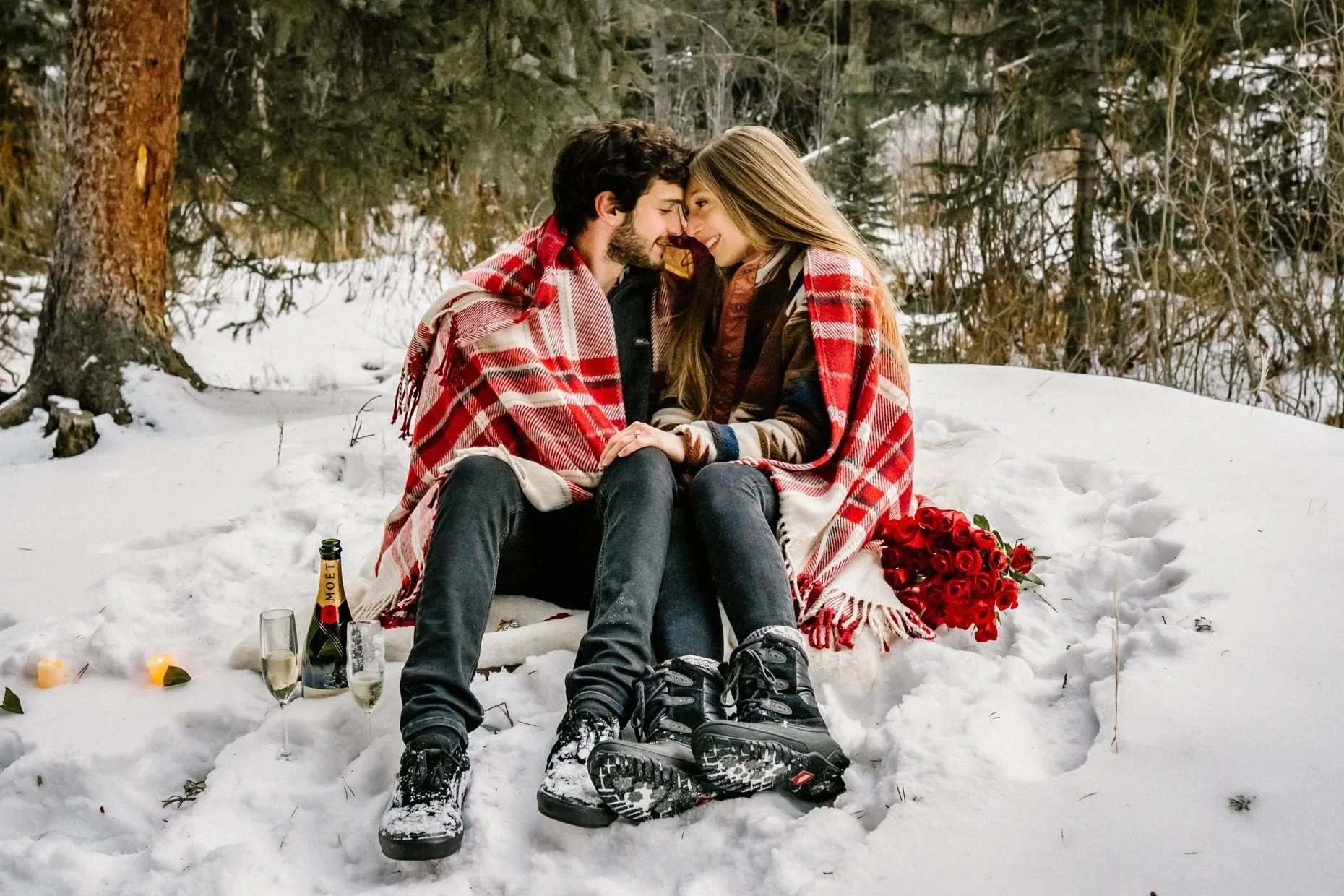 A man and women snuggle together under a flannel blanket while siting in the snow and her engagement ring is featured.