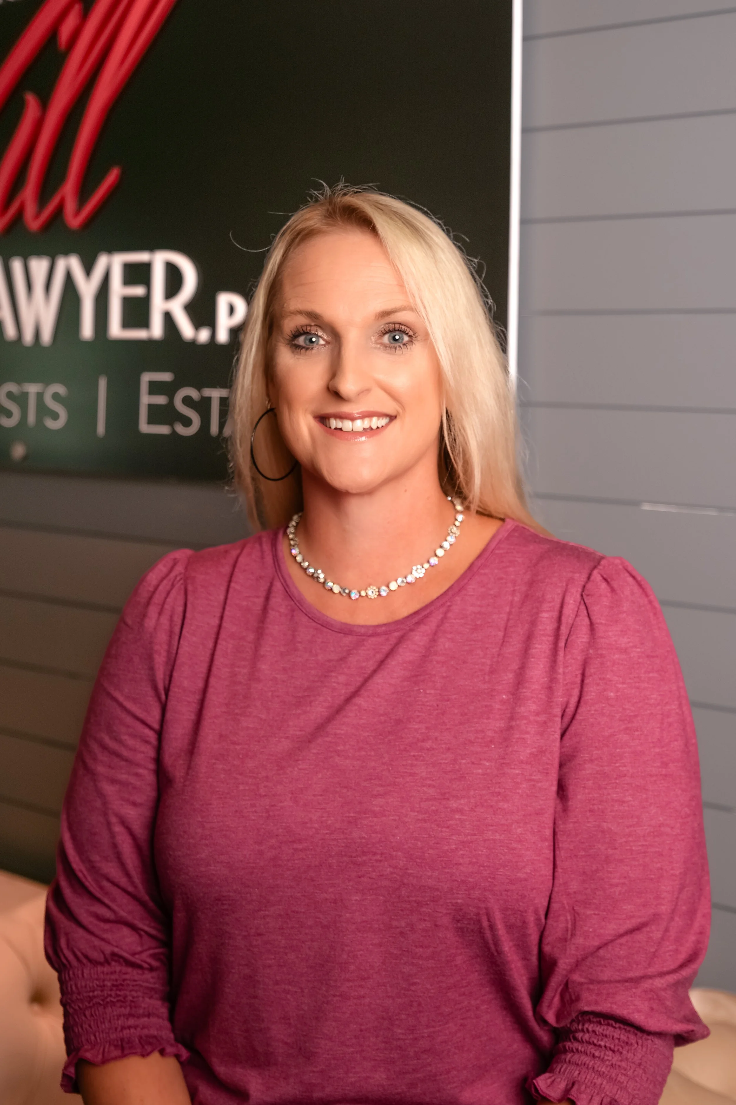 A woman with blonde hair wearing a pink top and pearl necklace, standing in front of a green sign with white text.
