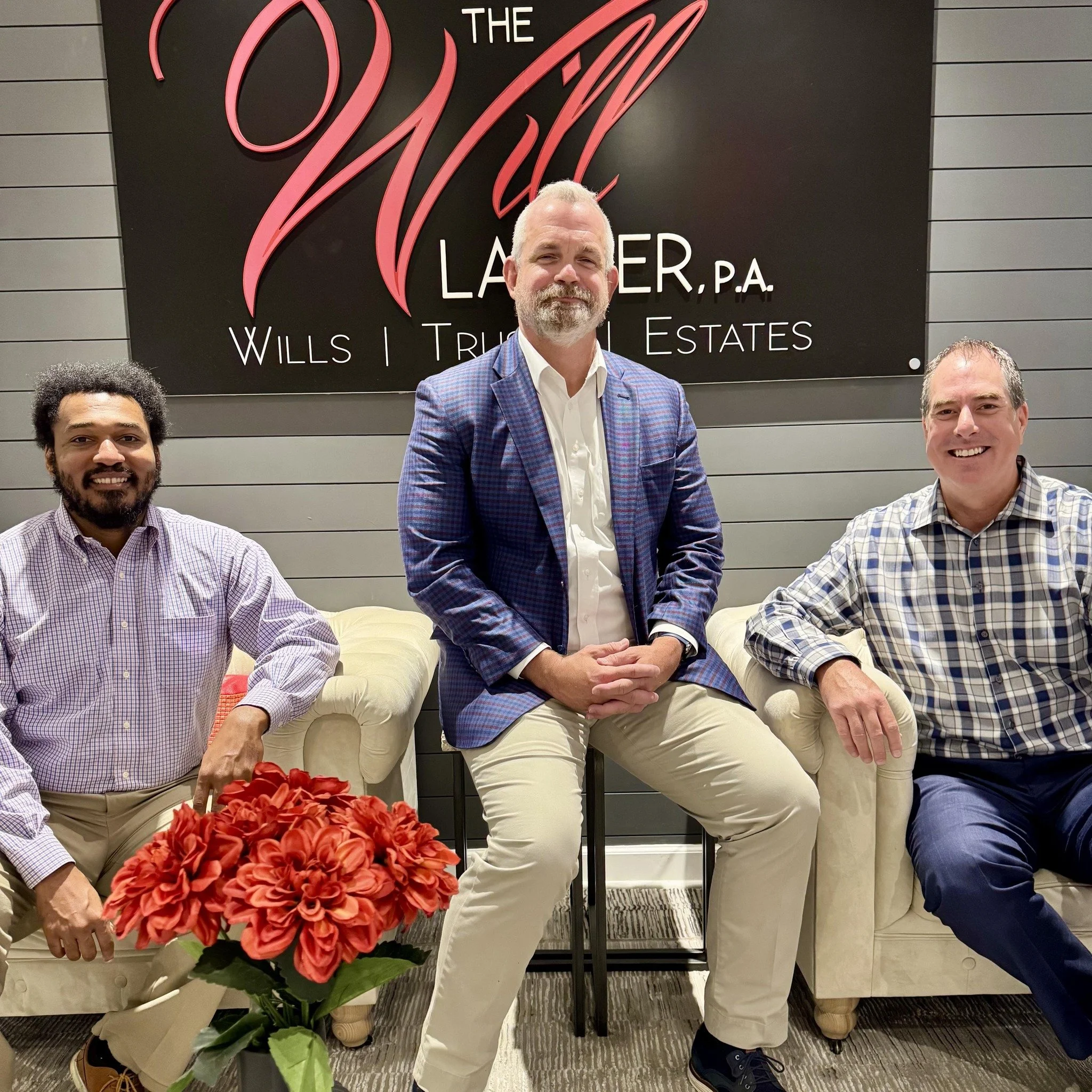 Three men sitting on a cream-colored couch in front of a sign that reads 'The Wills Law Firm, P.A., Wills, Trusts, Estates'. One man is in a plaid shirt, another in a patterned blazer, and the third in a checkered shirt. A bouquet of red flowers is on a small table in front of them.