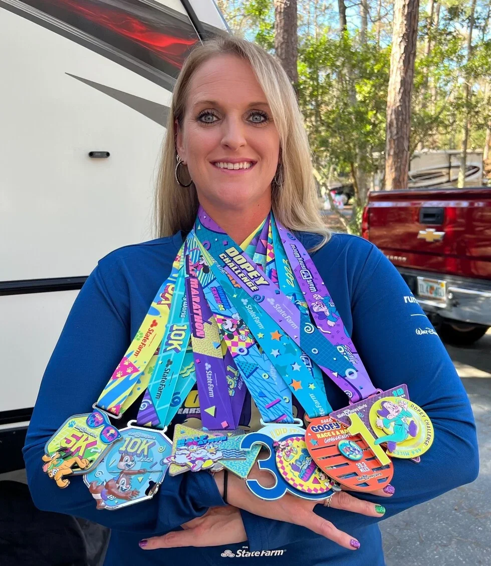 A woman wearing a blue long-sleeve shirt is smiling and holding numerous colorful race medals with Disney-themed designs, standing outdoors with trees and cars in the background.