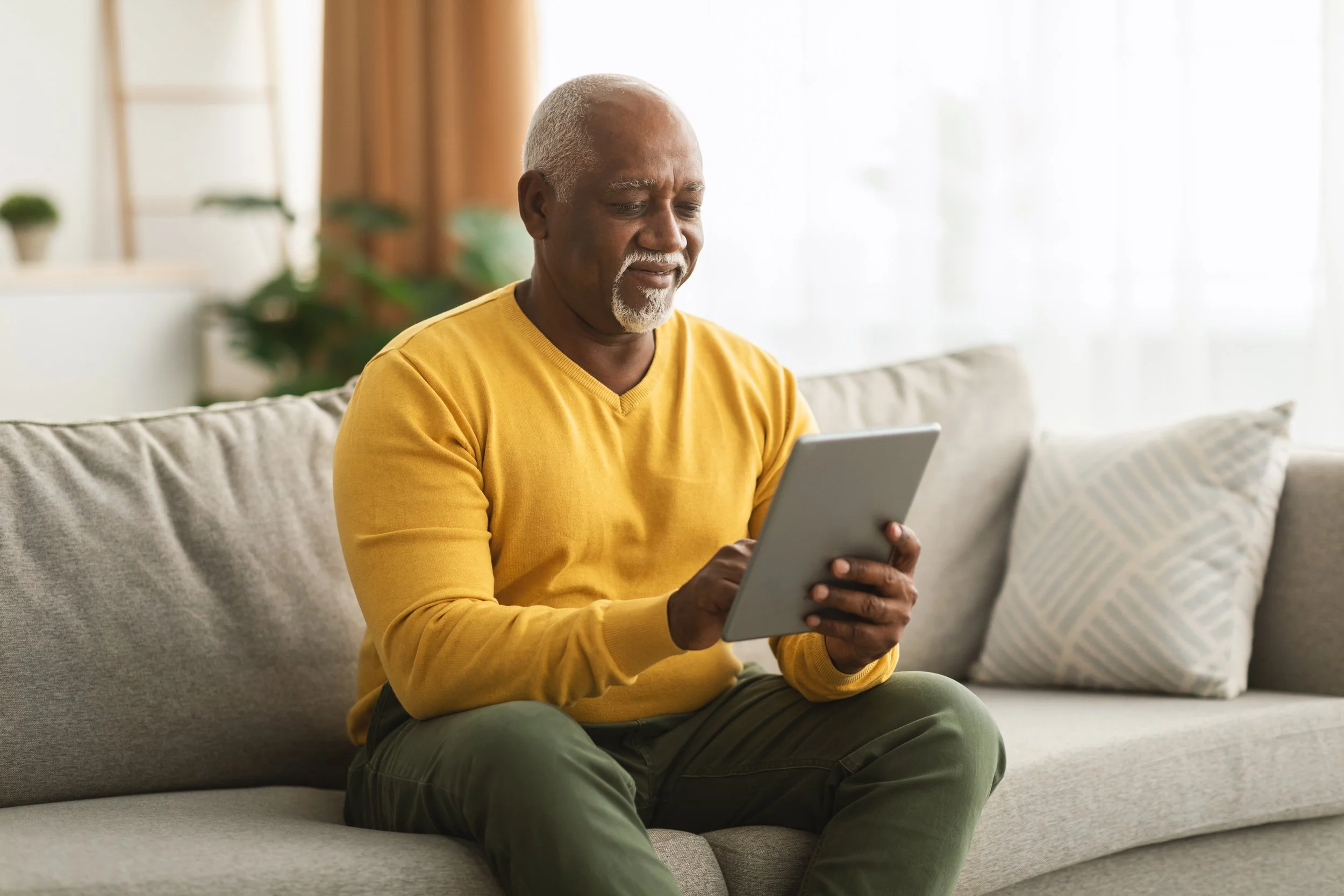 Older man sitting on a beige sofa, looking at a tablet in a bright living room with cushions and houseplants.
