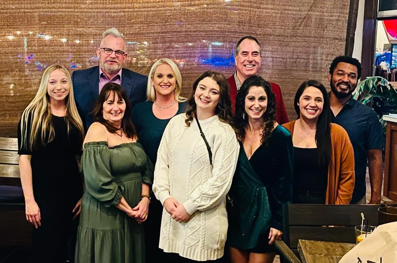 Group of ten people posing together at an indoor event, standing in front of a wooden wall, smiling and dressed in casual to semi-formal clothing.