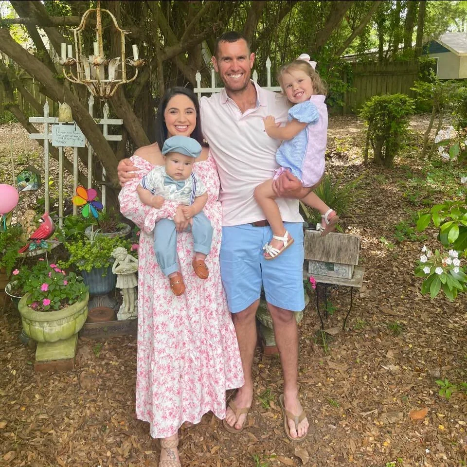 A family of four stands together outdoors in a garden, smiling at the camera. The woman is holding a baby boy, and the man is holding a young girl. The background features trees, plants, and garden decor like colorful pinwheels, a pink flamingo, and a chandelier hanging from a tree.
