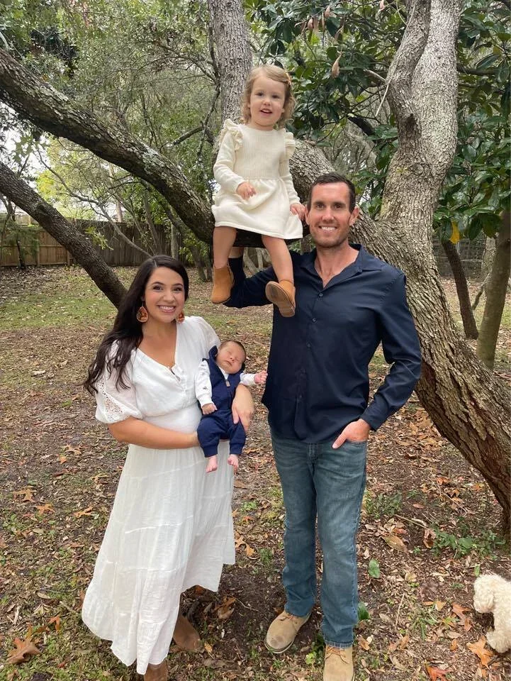 A family of four outdoors in a wooded area, with a woman holding a baby, a man holding a small girl sitting on a tree branch, and a dog partially visible on the ground.
