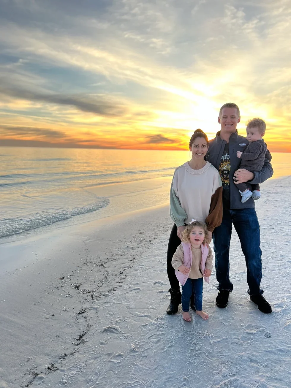 A family of four standing on a sandy beach at sunset, smiling and looking at the camera. The father is holding a young boy, the mother is standing beside him, and a young girl is standing in front of them. The sky is filled with colorful clouds and the ocean is in the background.