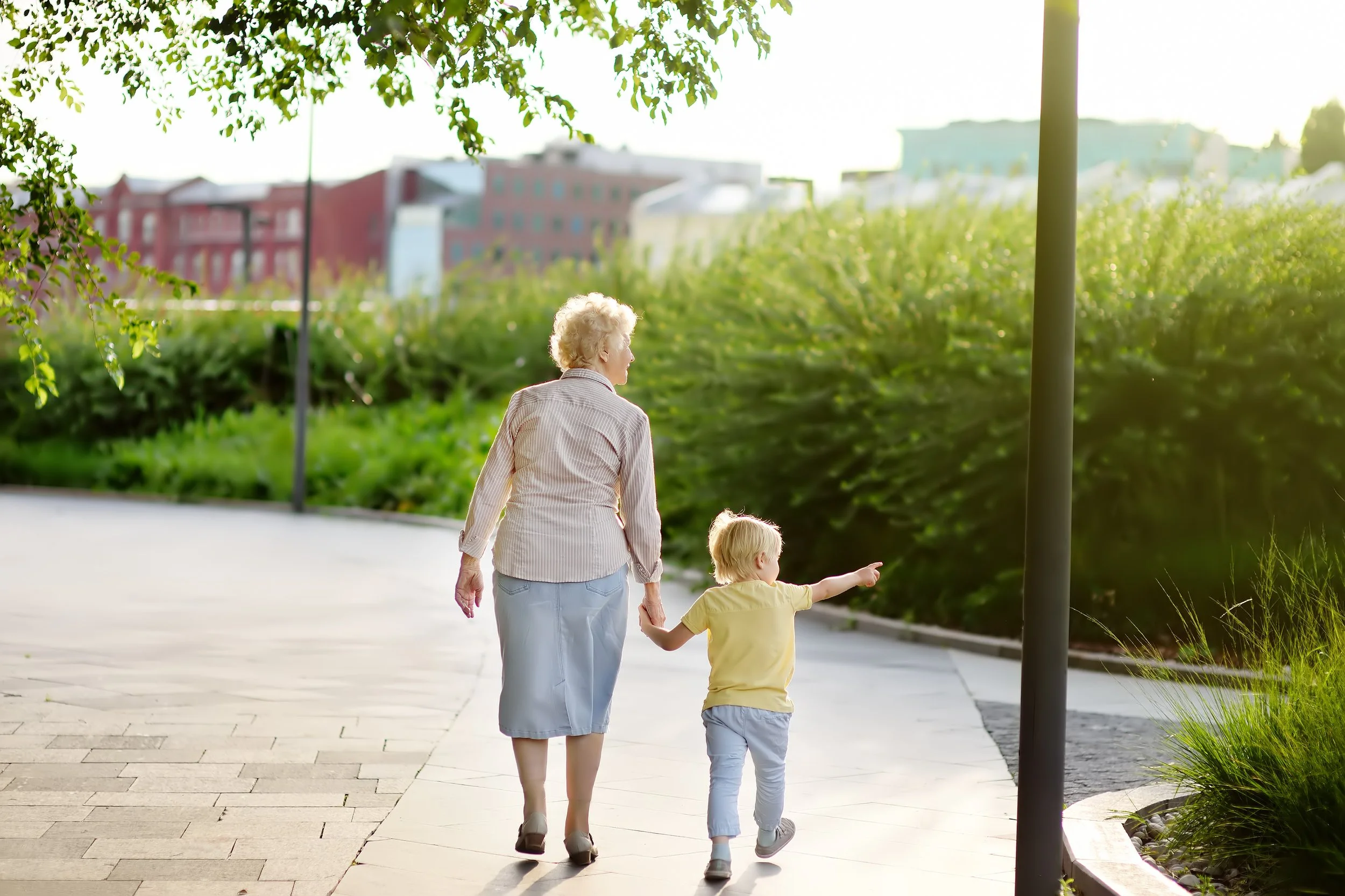 An elderly woman and a young boy walking hand in hand on a sidewalk in a park during daytime, with green bushes and buildings in the background.