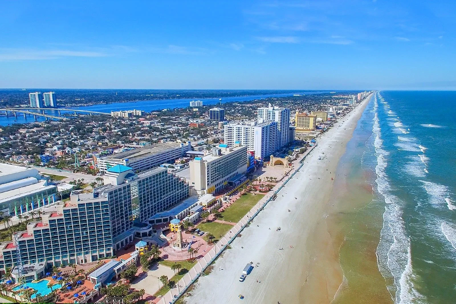 Aerial view of a coastal city with a long sandy beach, high-rise hotels, and a cityscape extending into the distance under a clear blue sky.