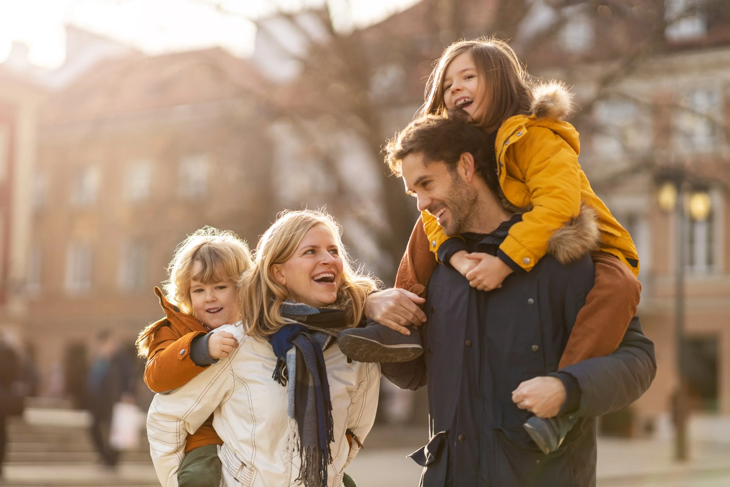 A happy family enjoying a day outdoors, with a man carrying a young girl on his shoulders and a woman walking beside him, with another young girl smiling nearby, in a park setting.