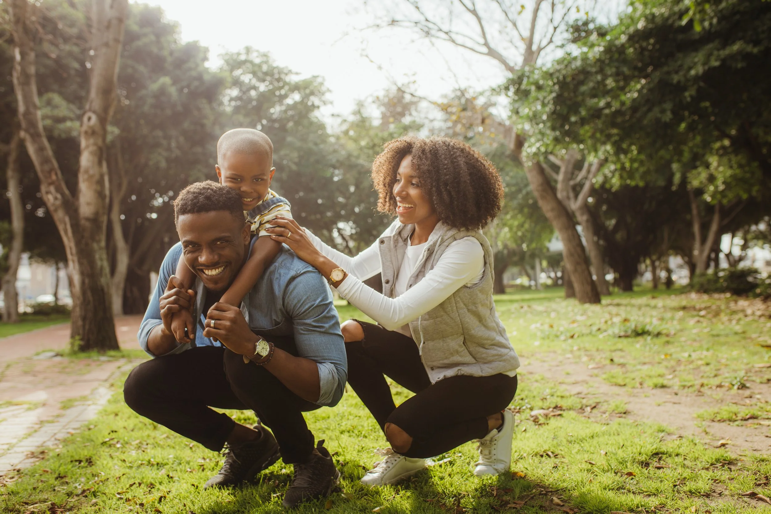 A happy family of three enjoying time in a park with tall trees, with a man crouching on the grass holding a young boy on his shoulders, and a woman kneeling beside them, all smiling.