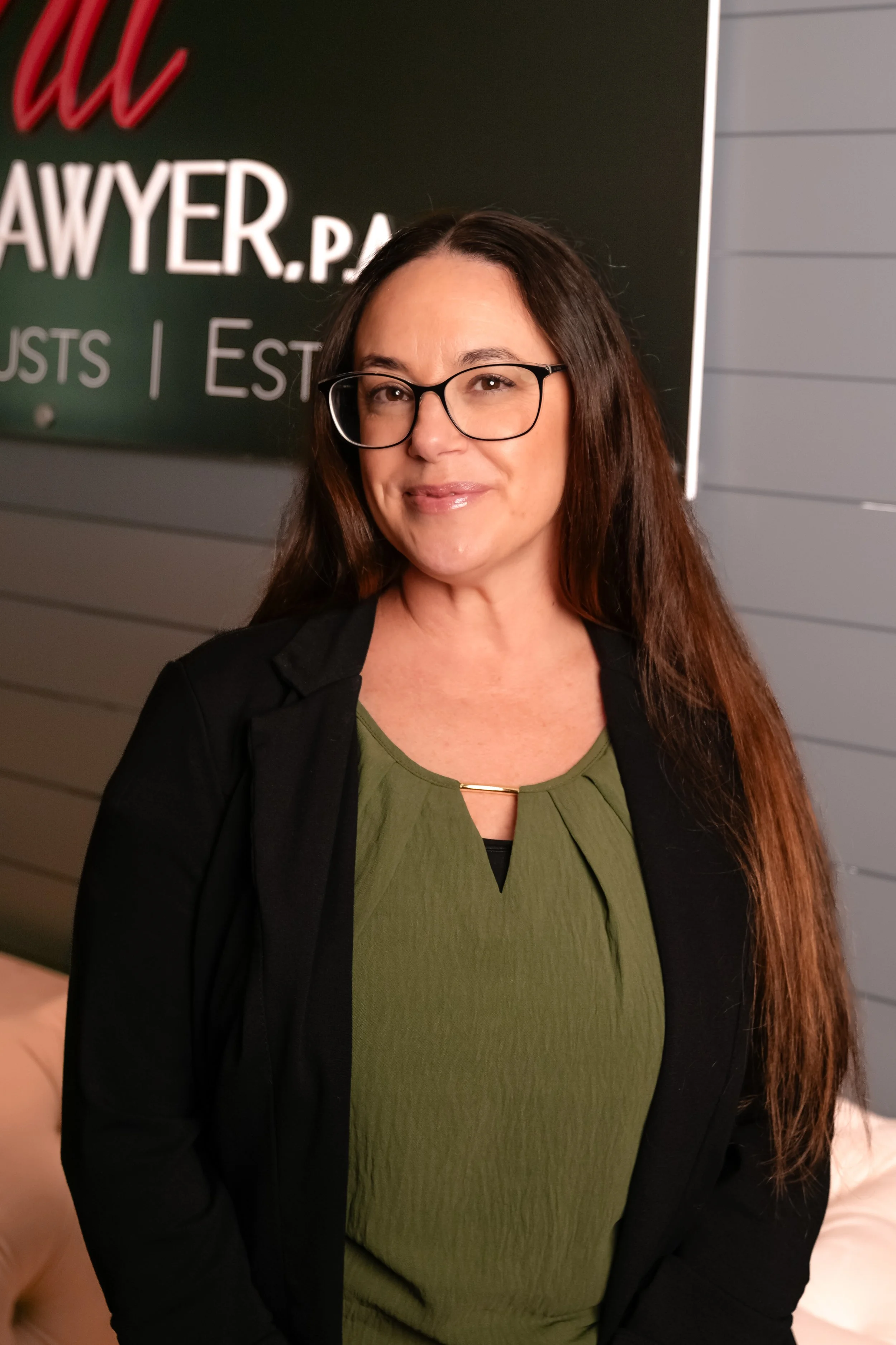 Woman with long brown hair and glasses, wearing a green top and black blazer, standing indoors in front of a gray wall with white stripes.