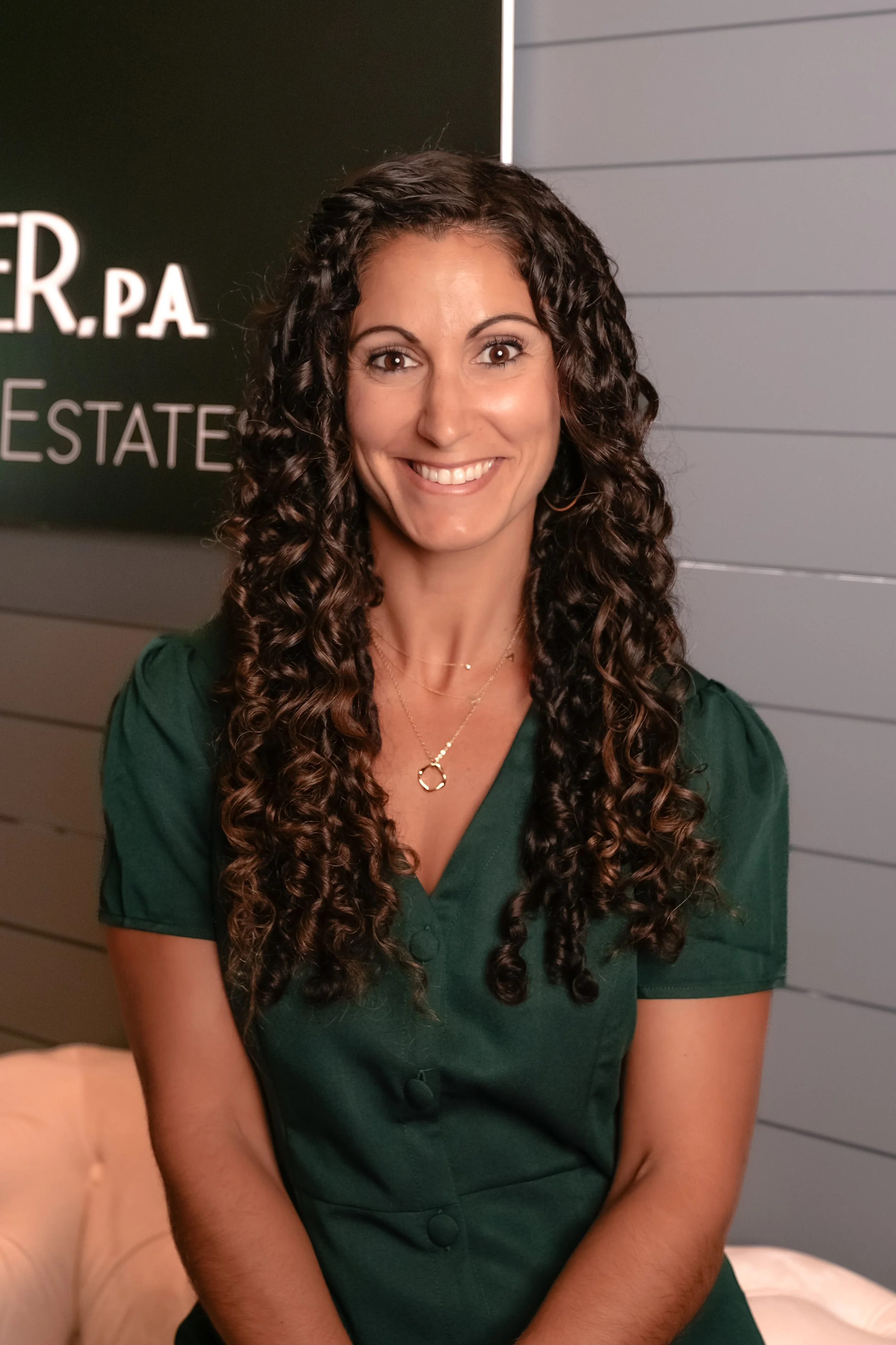 A smiling woman with long curly hair, wearing a green top, standing in front of a sign that says 'PA REAL ESTATE'.