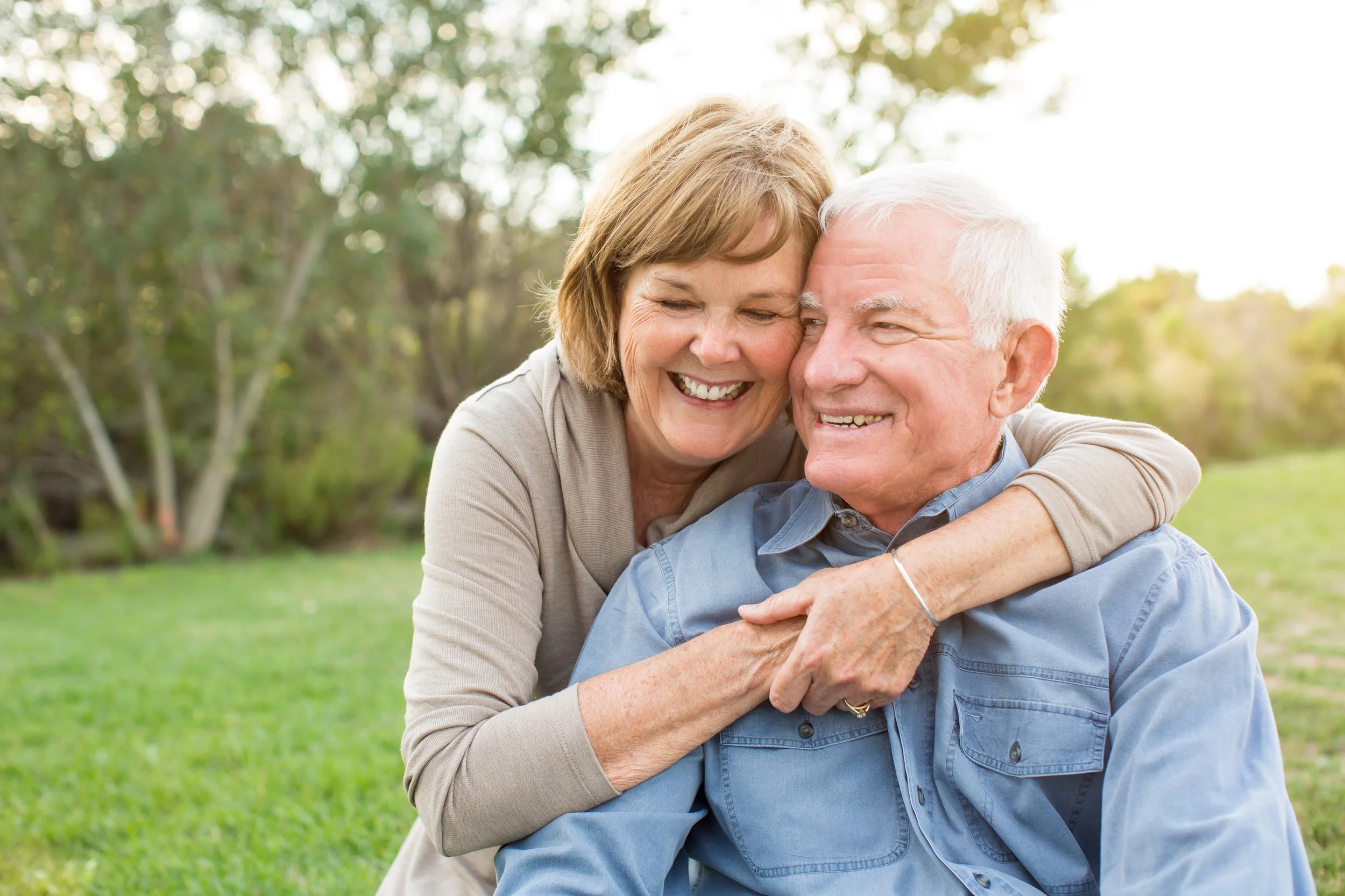 An elderly couple smiling and hugging outdoors in a park during daytime.