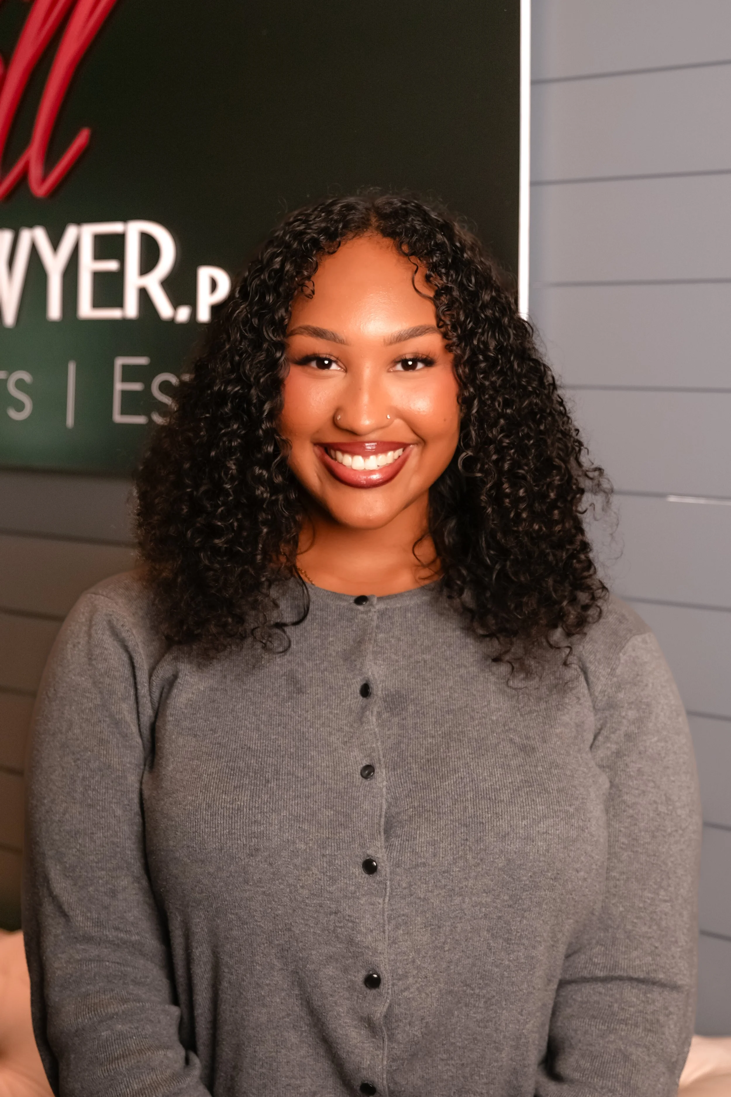 A young woman with curly black hair smiling in front of a gray wall background.