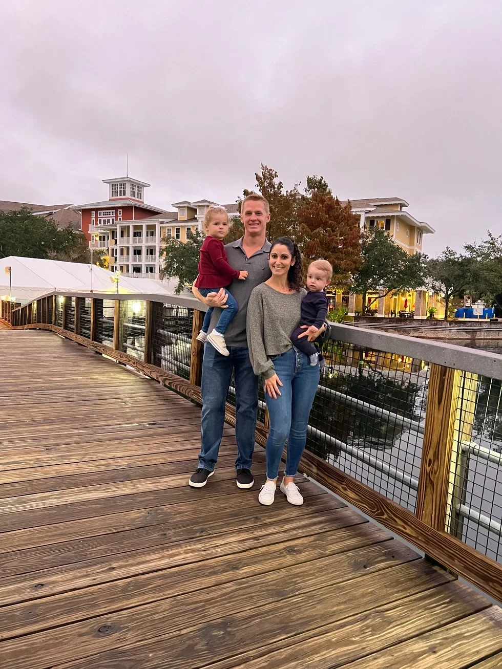 A family of four standing on a wooden bridge with water below, lined with trees and buildings in the background during overcast weather.