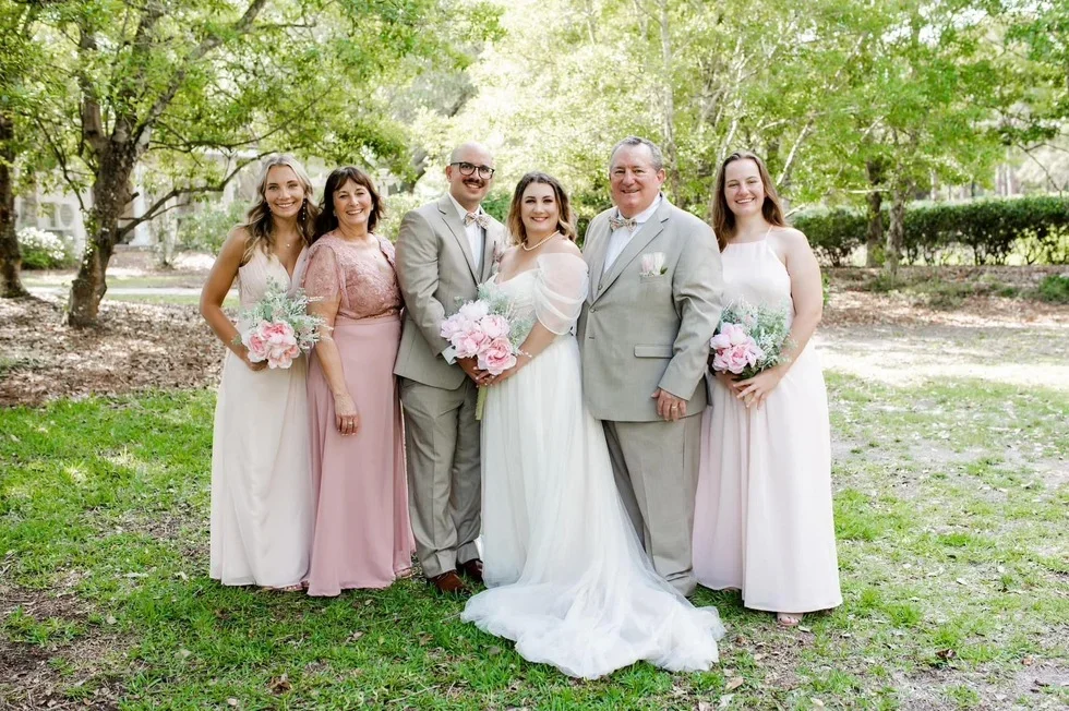 A wedding party standing outdoors on a lawn surrounded by trees. There are three women and three men, with the bride and groom in the center. The women are holding bouquets of pink and white flowers, and everyone is smiling.