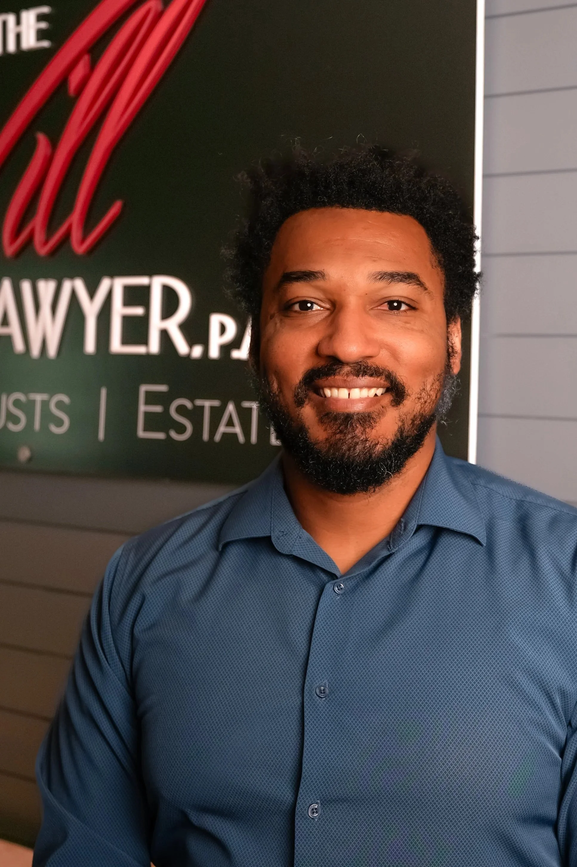 A man with a beard in a dark suit and light blue shirt standing indoors near a wall with a sign that reads "LAWYER" behind him.
