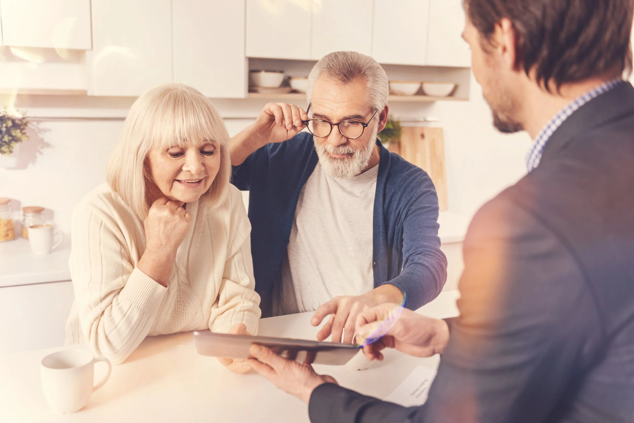 Three adults, two men and one woman, are gathered around a kitchen table looking at a tablet. The woman has white hair and is smiling, the man with glasses is adjusting his glasses, and the third man is holding the tablet.