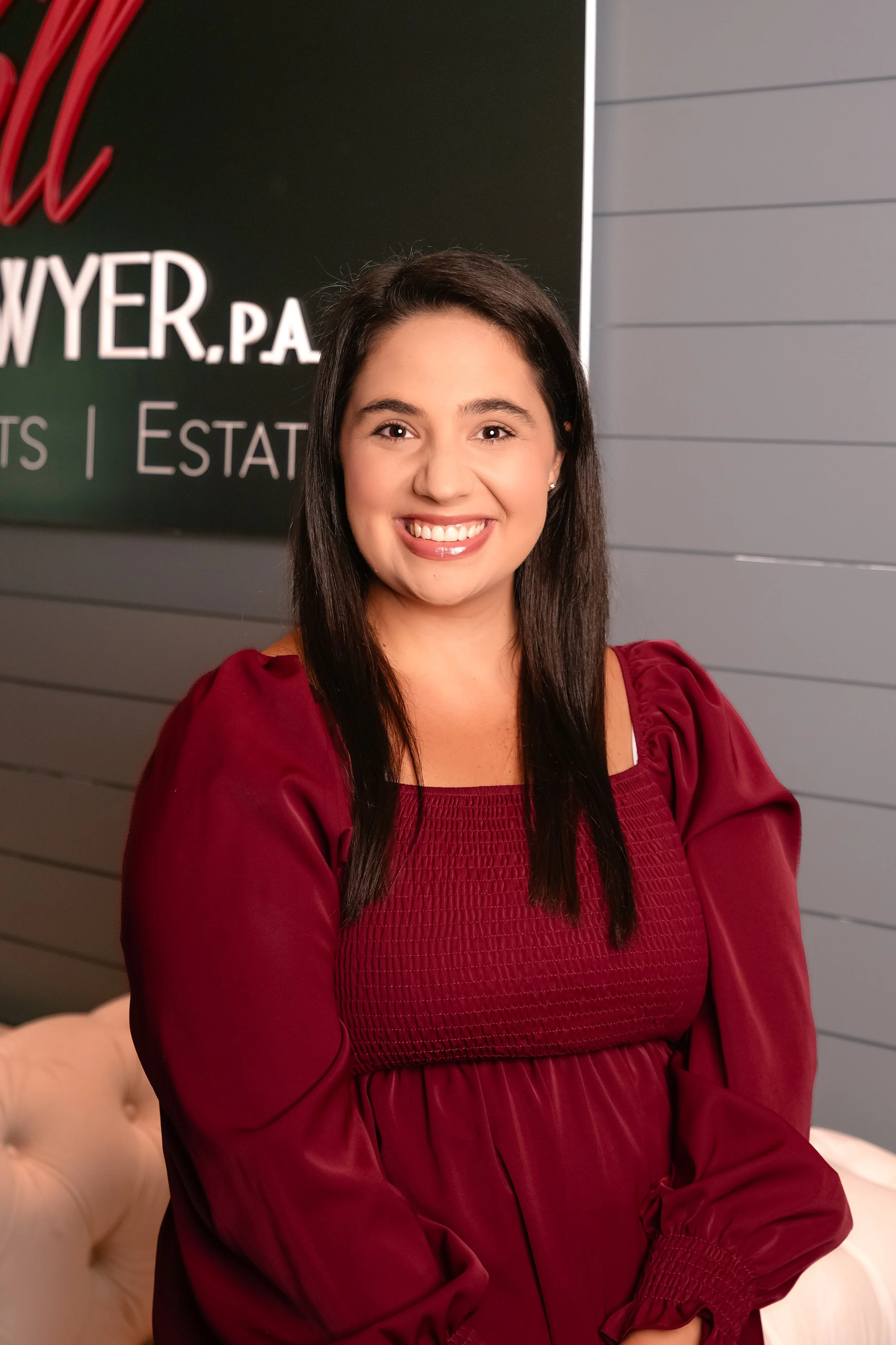 Smiling woman with long dark hair wearing a maroon top standing indoors against a gray wall with partial text.