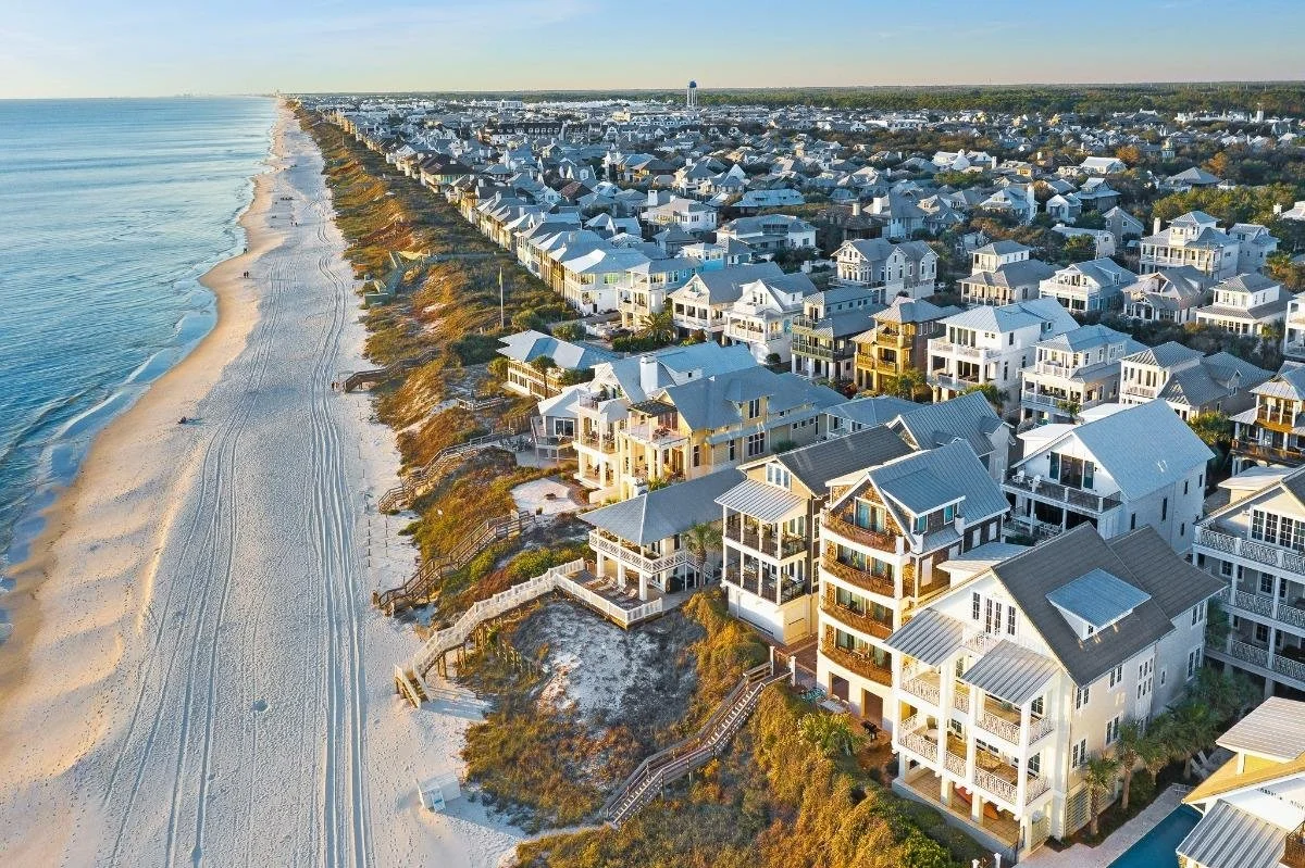 An aerial view of a coastal neighborhood with multiple white and light-colored beach houses along the shoreline, sandy beach with tire marks, and calm ocean waves on the left.