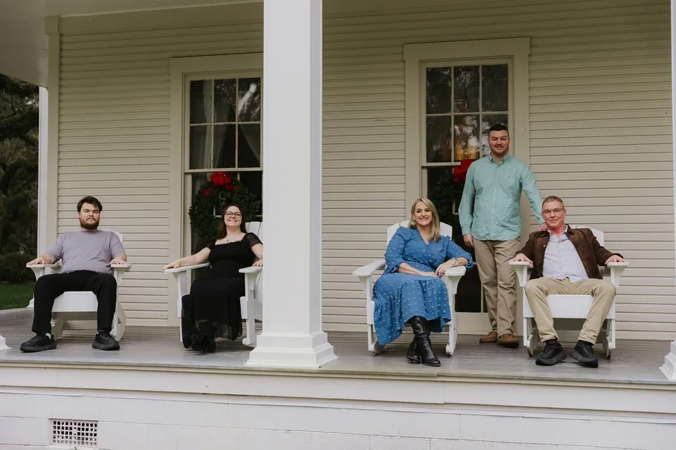 Four people sitting on porch chairs outside a house, with two additional people standing behind the seated individuals.