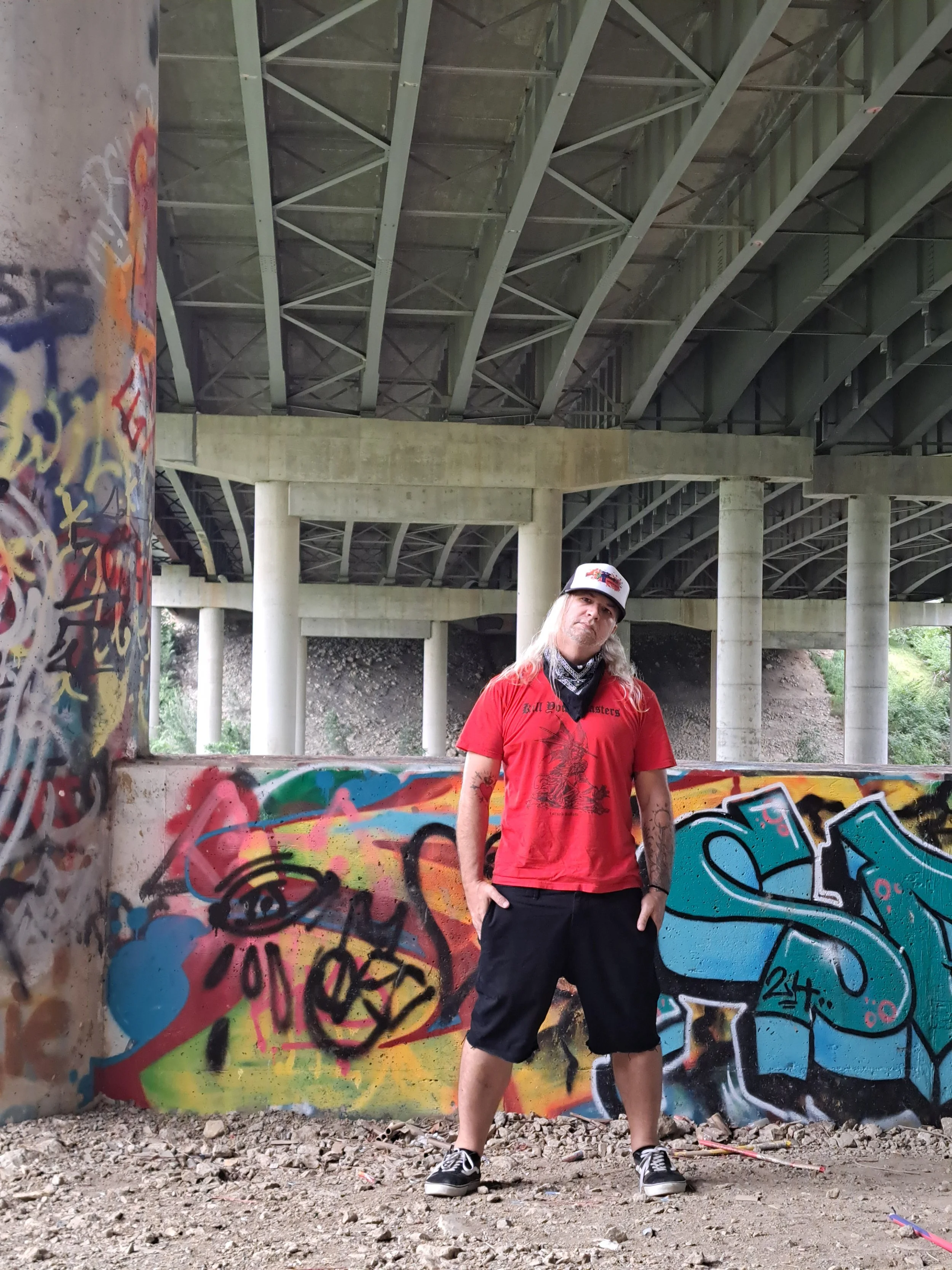 A man with long blonde hair wearing a red T-shirt, black shorts, and a trucker hat stands with hands in pockets under a bridge with graffiti on the wall behind him.