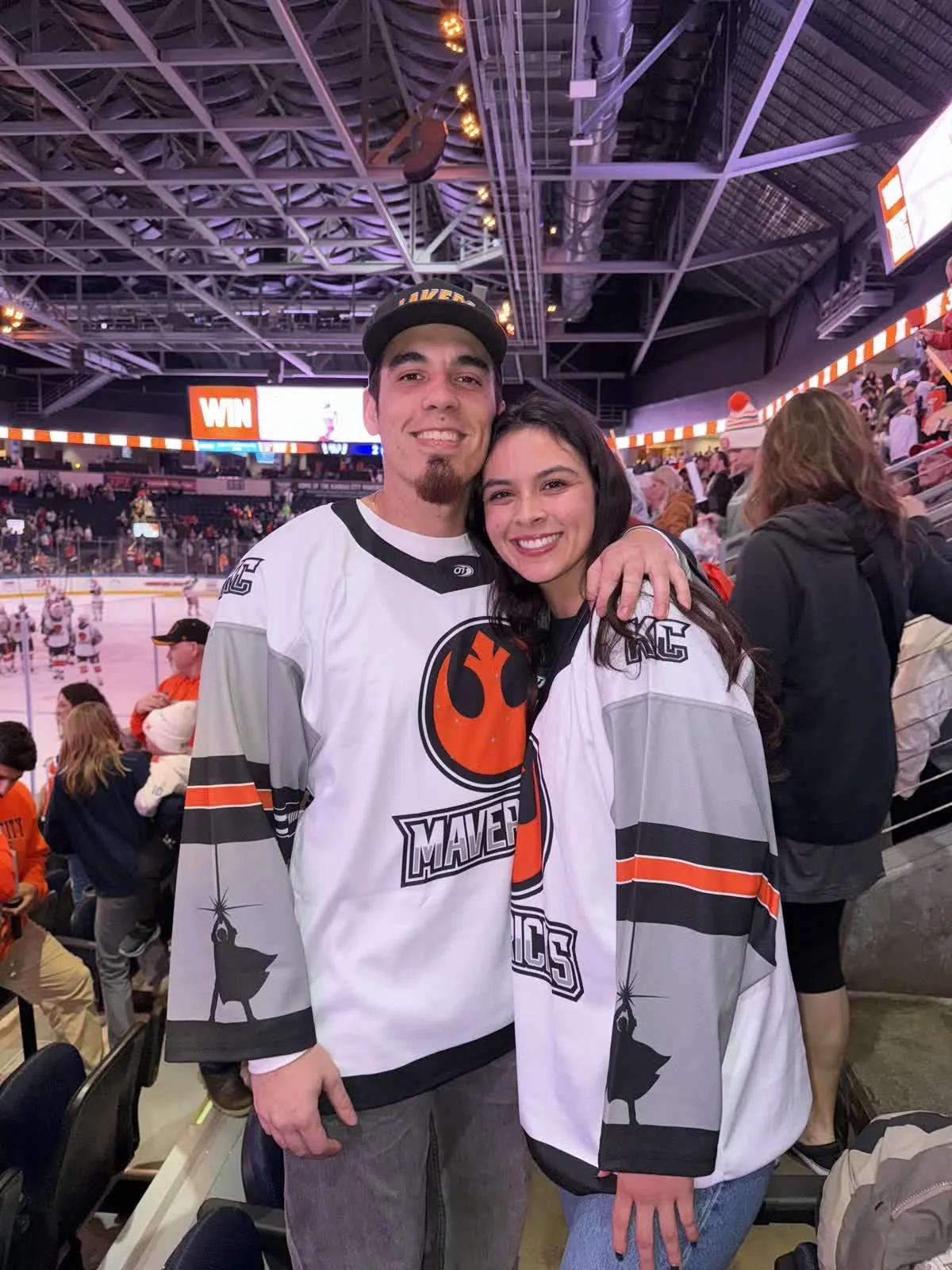Two people wearing hockey jerseys celebrating at an indoor ice hockey arena. The man has a beard and is wearing a black baseball cap. The woman has black hair. Behind them, the ice rink and other spectators are visible.
