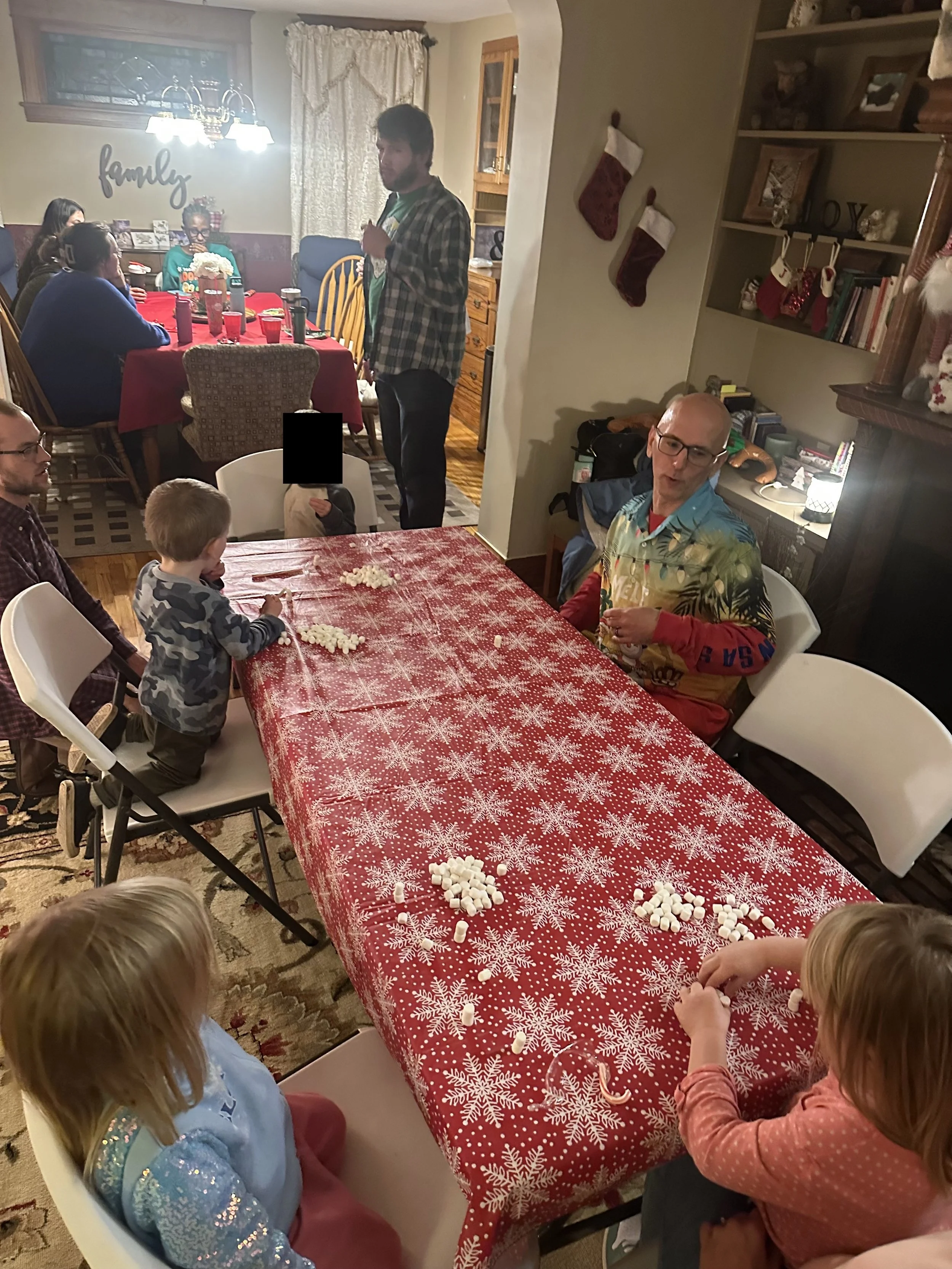 Family gathering around a table with a red snowflake-patterned tablecloth, children and adults celebrating, with Christmas stockings hanging on the wall, and holiday decorations in the background.