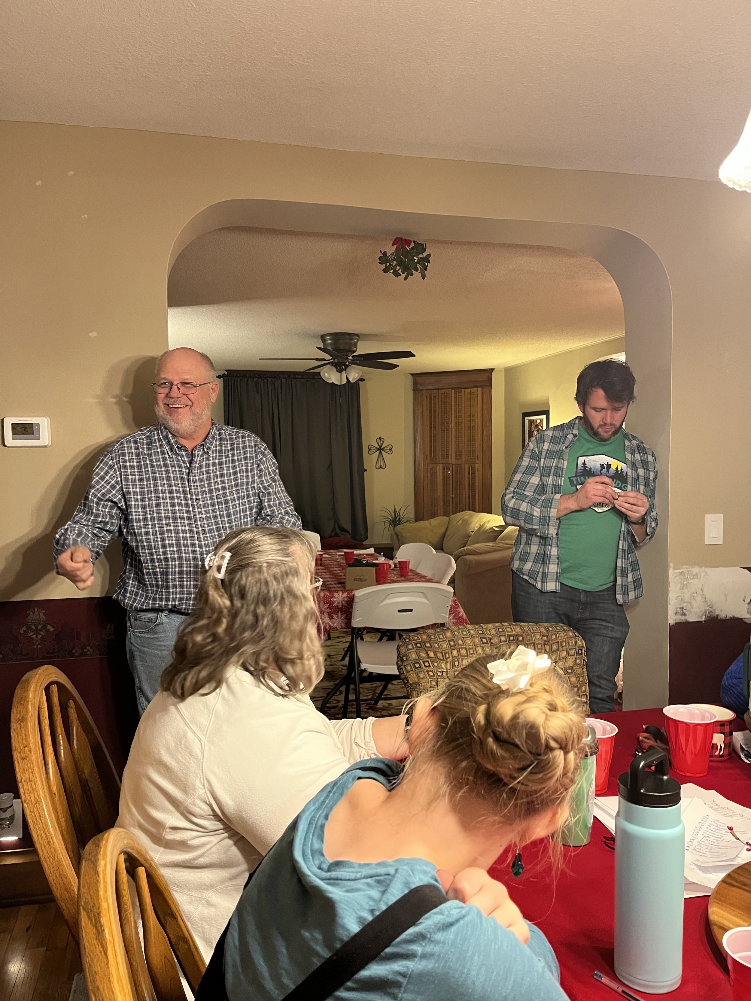 Family gathering around a dinner table in a cozy home, with a man smiling and a younger man looking at his phone, in a warmly lit room decorated for a holiday celebration.